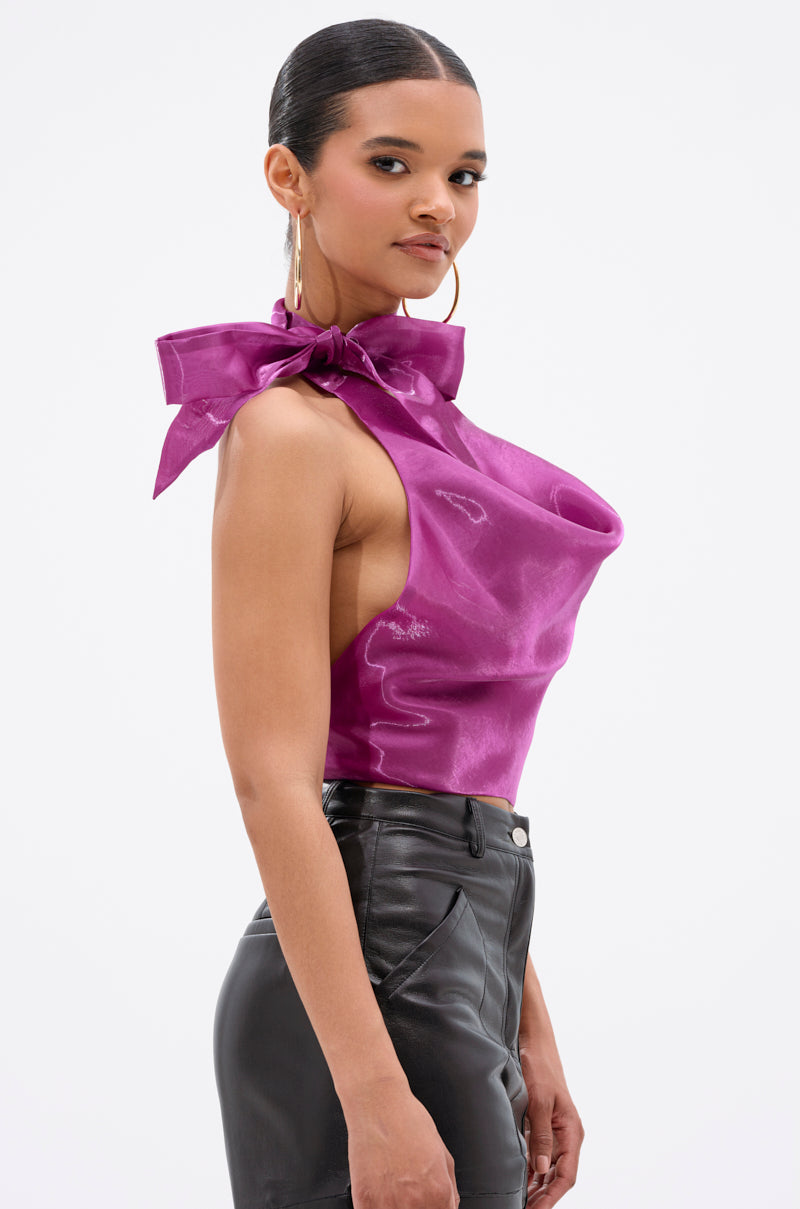 A woman poses sideways against a white background, wearing the SUNSHINE COWL NECK SATIN BLOUSE IN PURPLE, featuring a shiny finish, large bow, open-back design, and paired with high-waisted black leather pants. 3