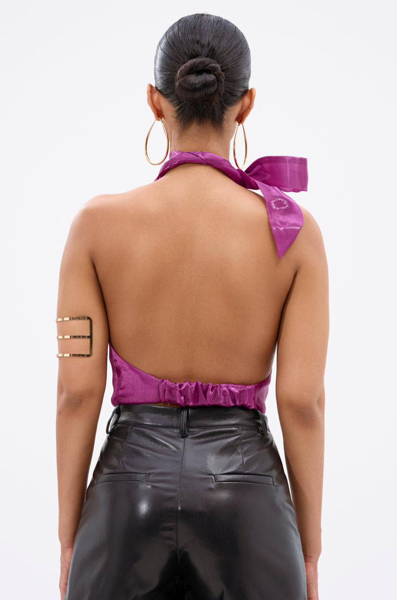 A person with their back to the camera wears the SUNSHINE COWL NECK SATIN BLOUSE IN PURPLE, featuring an open-back design, paired with black shiny pants, a neat bun, large hoop earrings, and three gold arm cuffs. 4