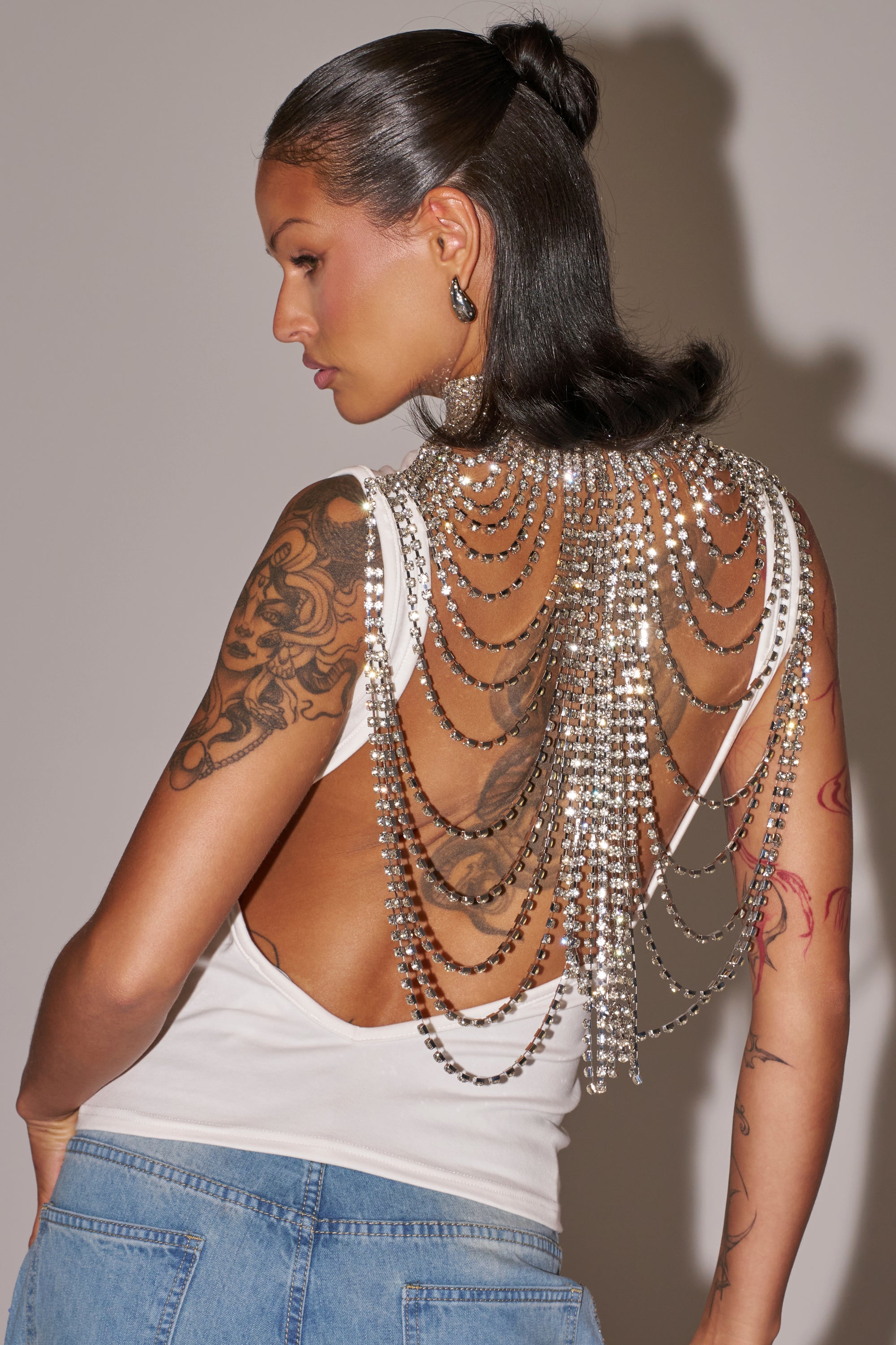 A tattooed person wears the ONE OF ONE RHINESTONE SLEEVELESS TEE—a backless top adorned with layered, sparkling chains and jewels—paired with slicked-back hair, silver earrings, and blue jeans for a bold festival look. 0