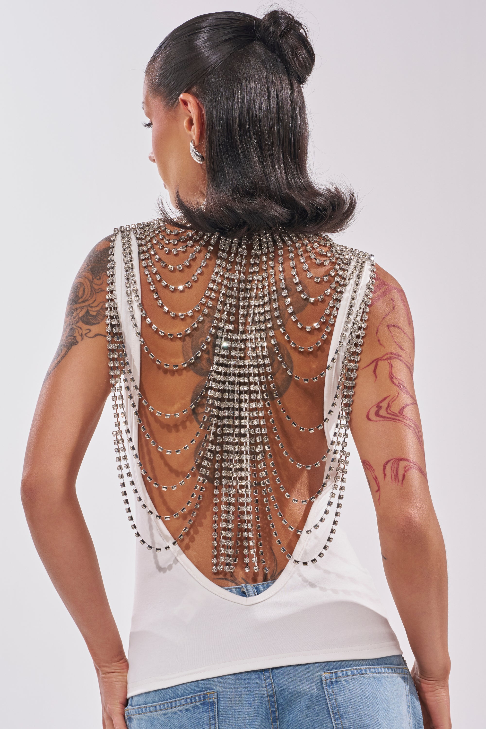 A person with dark, slicked-back hair wears the ONE OF ONE RHINESTONE SLEEVELESS TEE with cascading silver chains, paired with light blue jeans. Tattoos are visible on their arms. 3