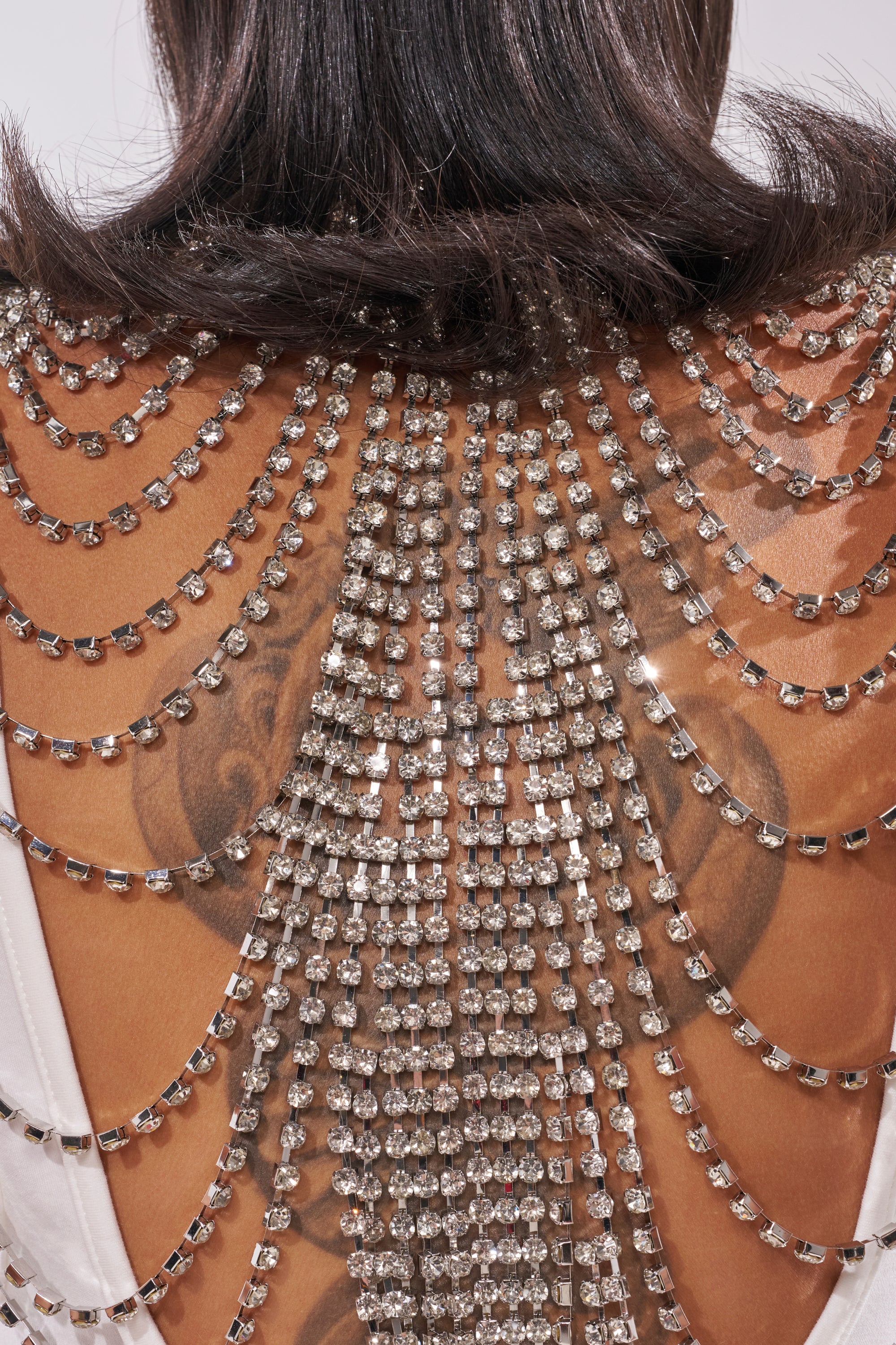 Close-up of a woman's back with long dark hair, wearing the ONE OF ONE RHINESTONE SLEEVELESS TEE featuring a rhinestone chain design on a backless white top—revealing tattoos underneath for a bold festival look. 8