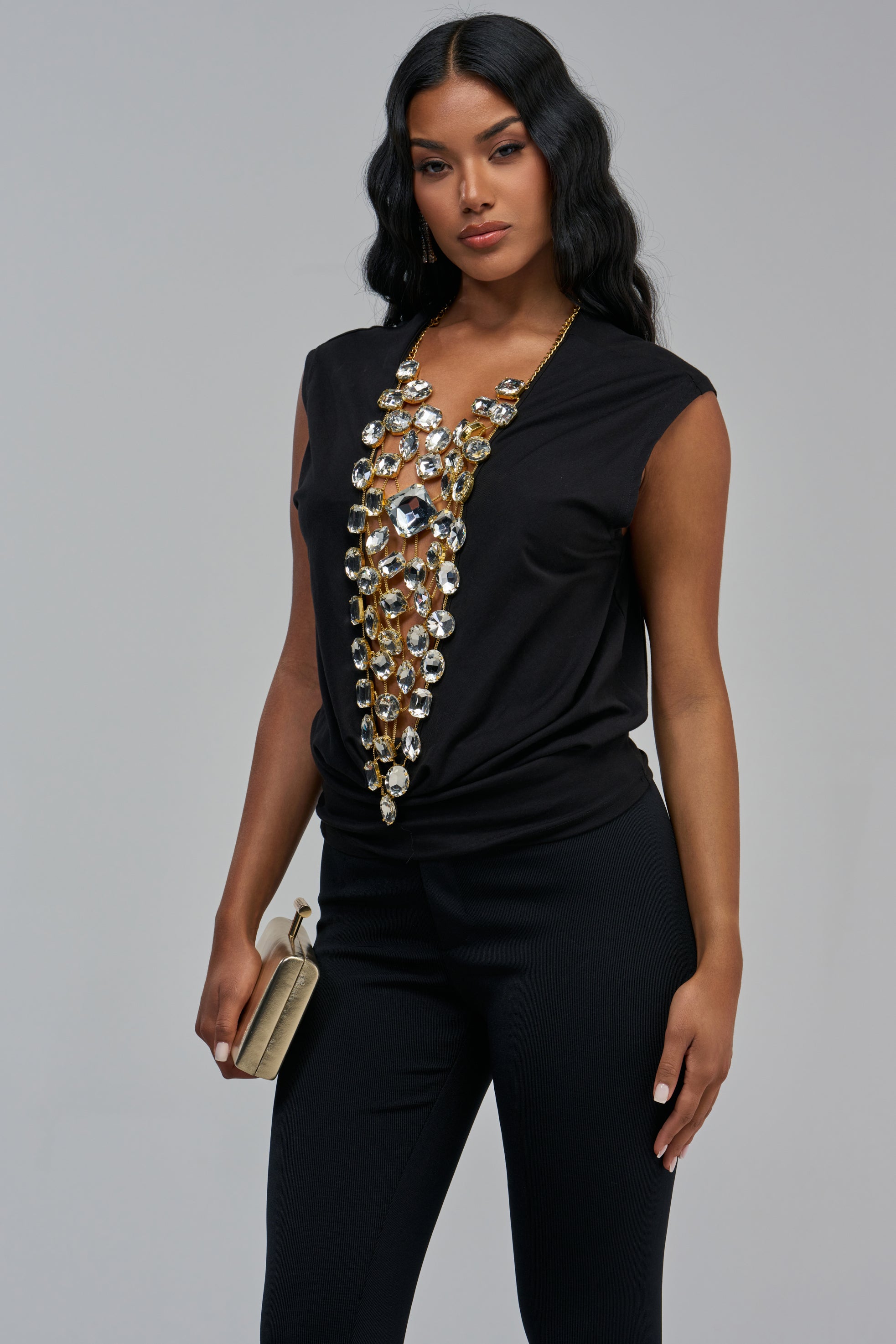 The LAYLA RHINESTONE TEE IN BLACK is a sleeveless tank top featuring a cascading gold chain necklace with large faceted rhinestones, worn with black trousers and a gold clutch.