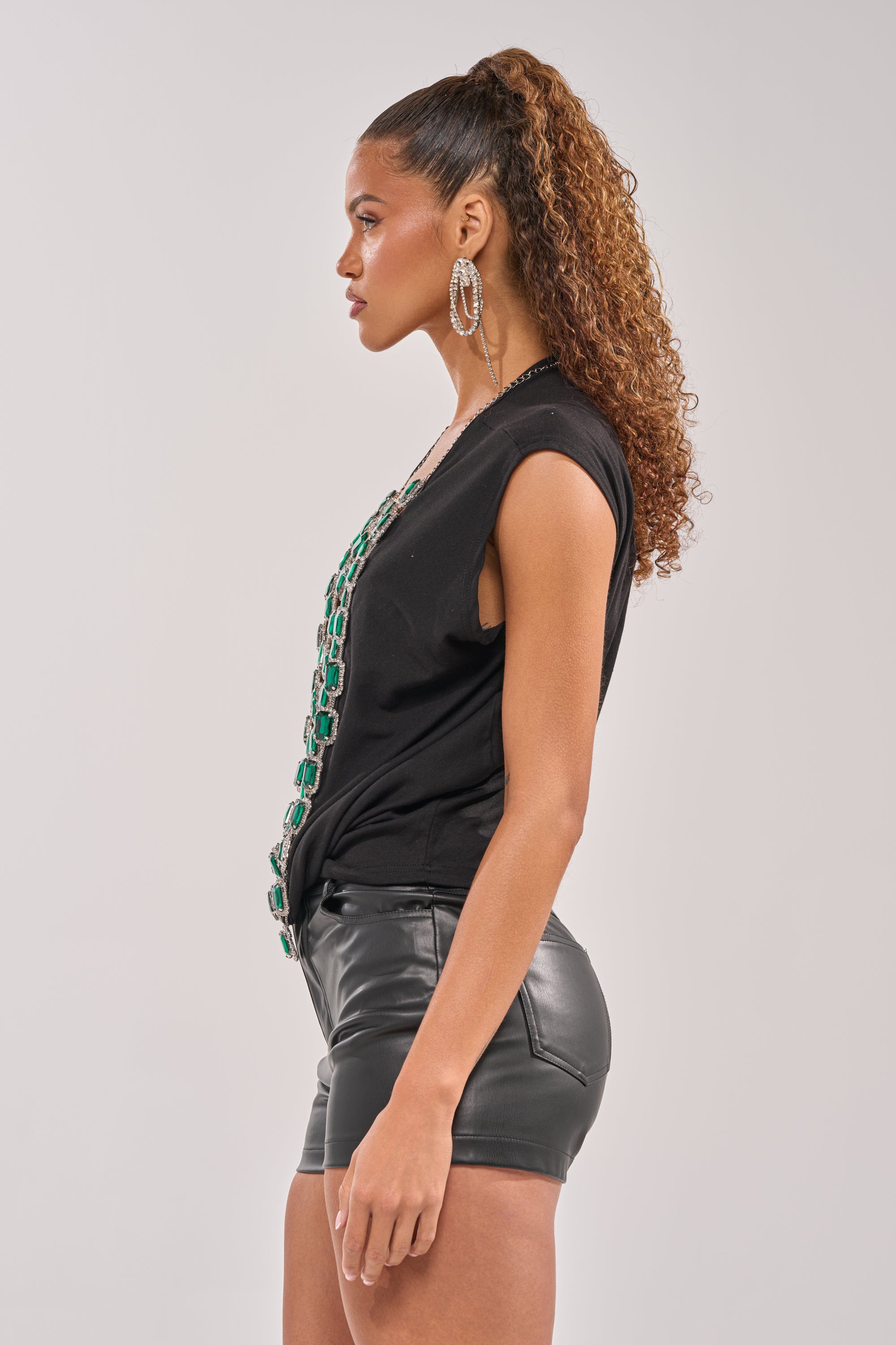 A woman with curly hair in a high ponytail stands in profile, wearing large hoop earrings, black leather shorts, and the EMERALD BABY RHINESTONE TEE—creating a bold streetwear look against a plain background. 3