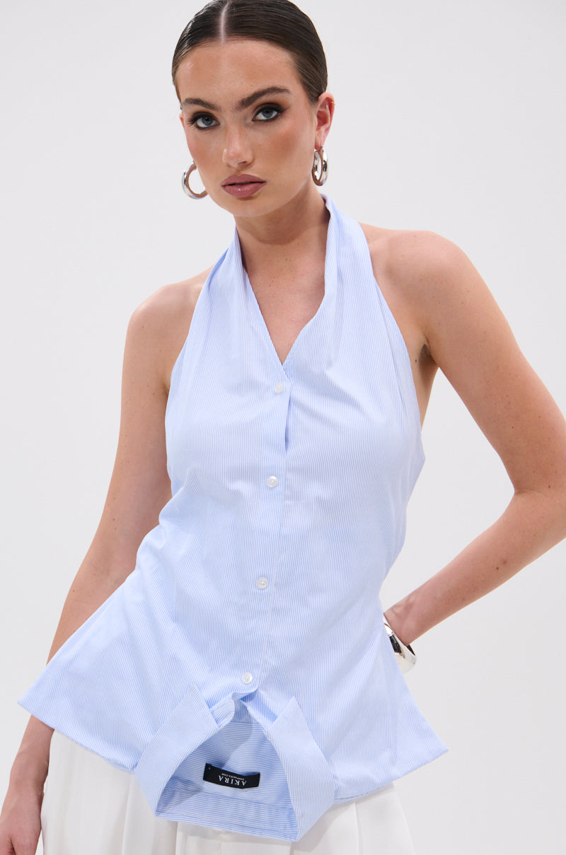 A woman with slicked-back hair wears the TINA STRIPE POPLIN BLOUSE—a halter-style, pinstriped light blue button-up with a flared hem—paired with white bottoms, posing confidently with one hand on her hip against a plain white background. 0