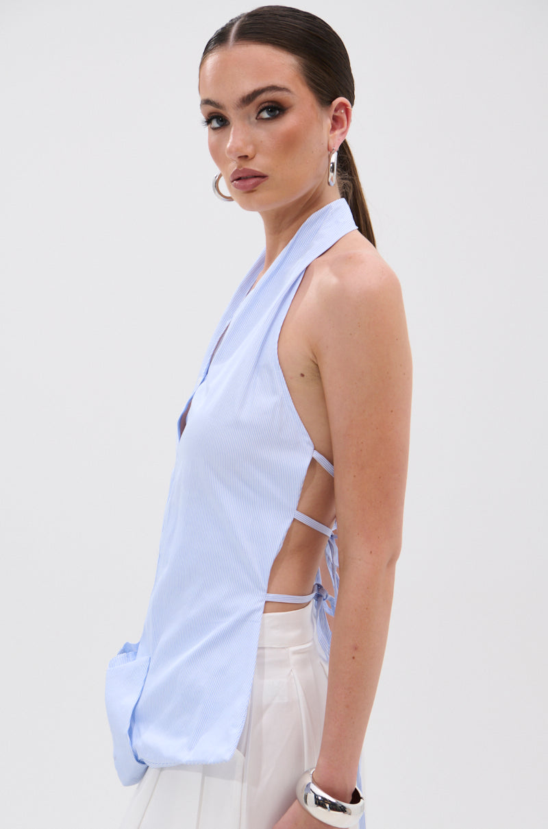 A woman models the TINA STRIPE POPLIN BLOUSE, a sleeveless halter-style top with side ties and a plunging neckline, paired with a sheer white skirt. She accessorizes with hoop earrings, a chunky bracelet, and slicked-back hair. 3