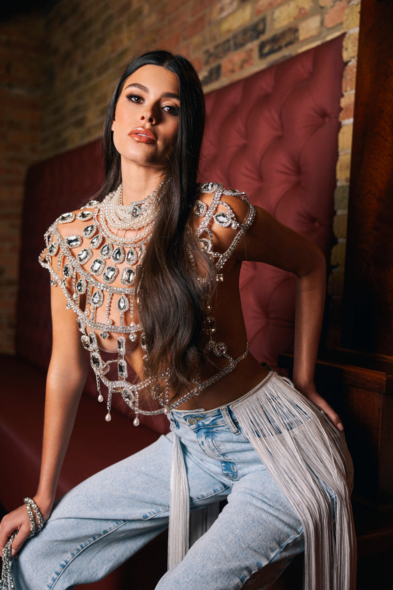 A woman with long dark hair sits on a maroon tufted bench against a brick wall, confidently posing in the RUBY QUEEN RHINESTONE BLOUSE—an eye-catching top with pearl-embellished details—paired with light blue fringe jeans.