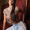 A woman with long dark hair sits on a maroon tufted bench against a brick wall, confidently posing in the RUBY QUEEN RHINESTONE BLOUSE—an eye-catching top with pearl-embellished details—paired with light blue fringe jeans.