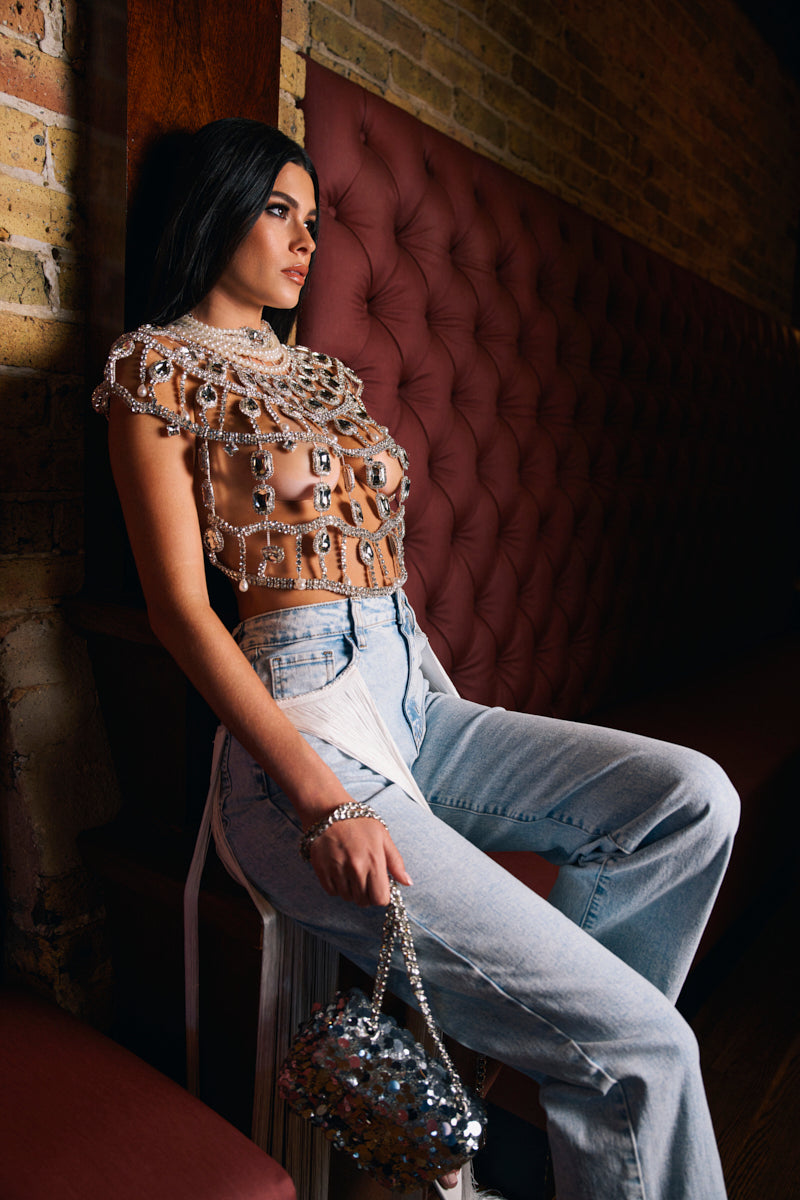 A woman with long dark hair sits on a bench against a brick wall and tufted maroon upholstery. She wears the RUBY QUEEN RHINESTONE BLOUSE, high-waisted light blue jeans, and holds a sparkling silver purse.