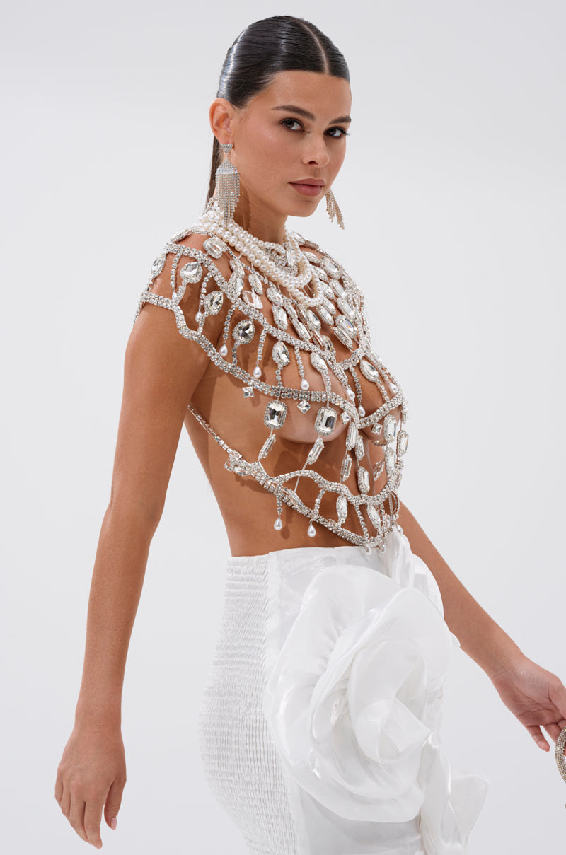 A woman poses in the RUBY QUEEN RHINESTONE BLOUSE with cutouts and crystal-cut gems, paired with a white textured skirt featuring a large fabric flower. Her hair is sleek and pulled-back, accessorized by dangling earrings.