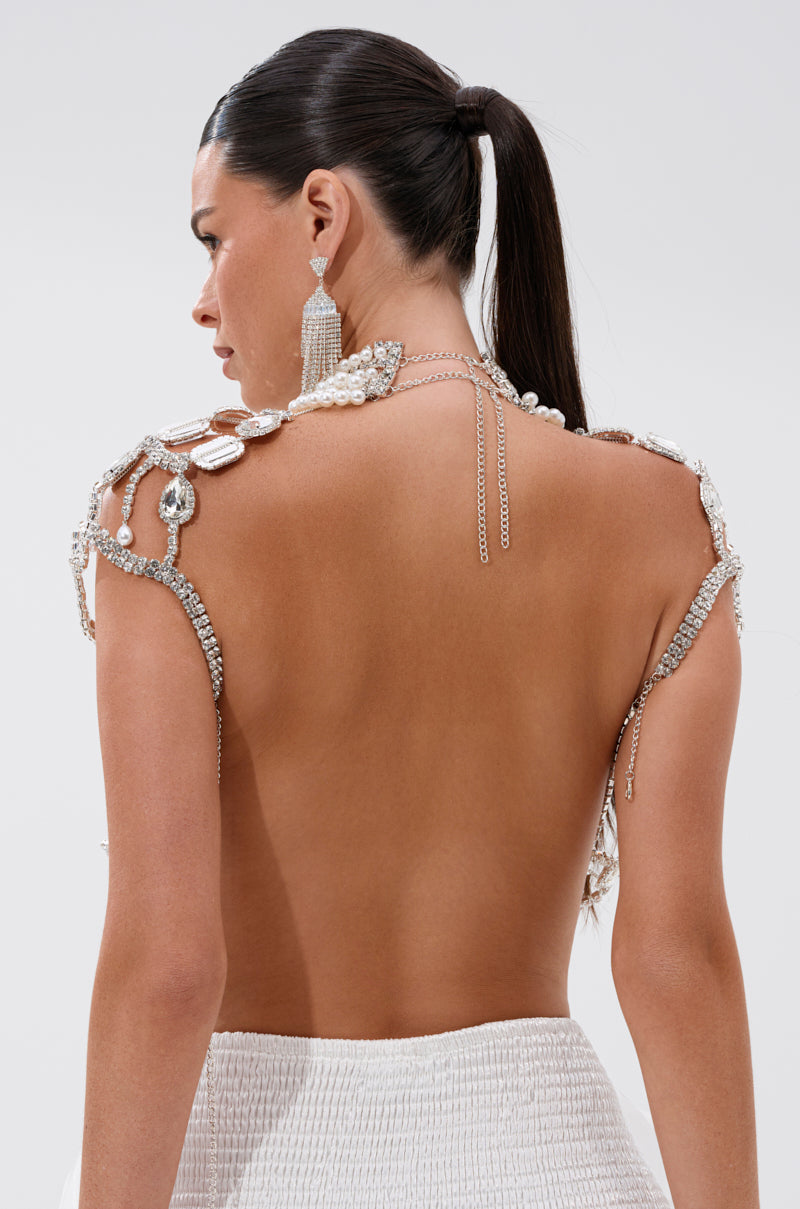 A woman with a sleek ponytail is shown from the back, wearing the RUBY QUEEN RHINESTONE BLOUSE, featuring metallic and pearl shoulder details, chainlink draping across her upper back, and a large tasseled earring.
