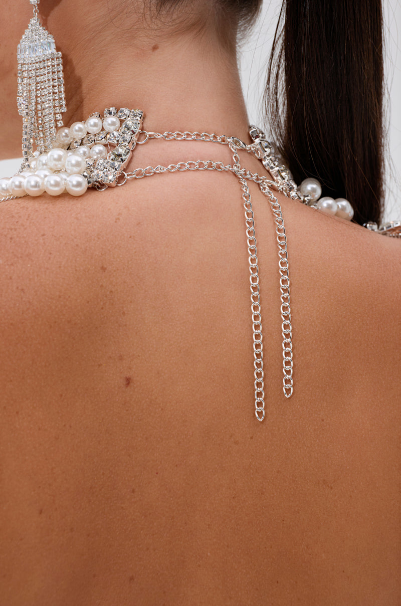 Close-up of a woman's upper back wearing the RUBY QUEEN RHINESTONE BLOUSE, featuring layered silver chains, pearl accents, and crystal-cut gems, with a glimpse of a dangling beaded earring and dark hair tied back.