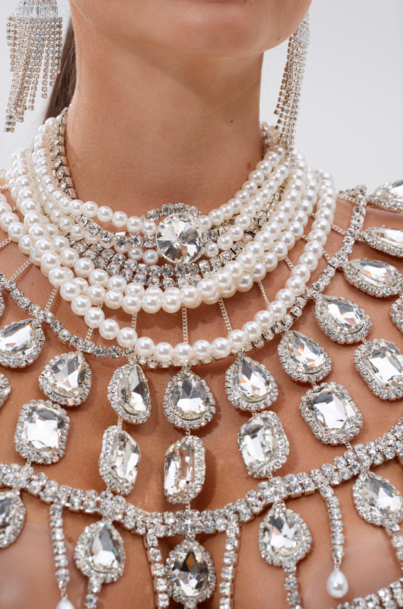 A close-up of a person wearing the RUBY QUEEN RHINESTONE BLOUSE, layered pearl necklaces, and matching chandelier earrings with large gemstones, creating a glamorous and luxurious look.