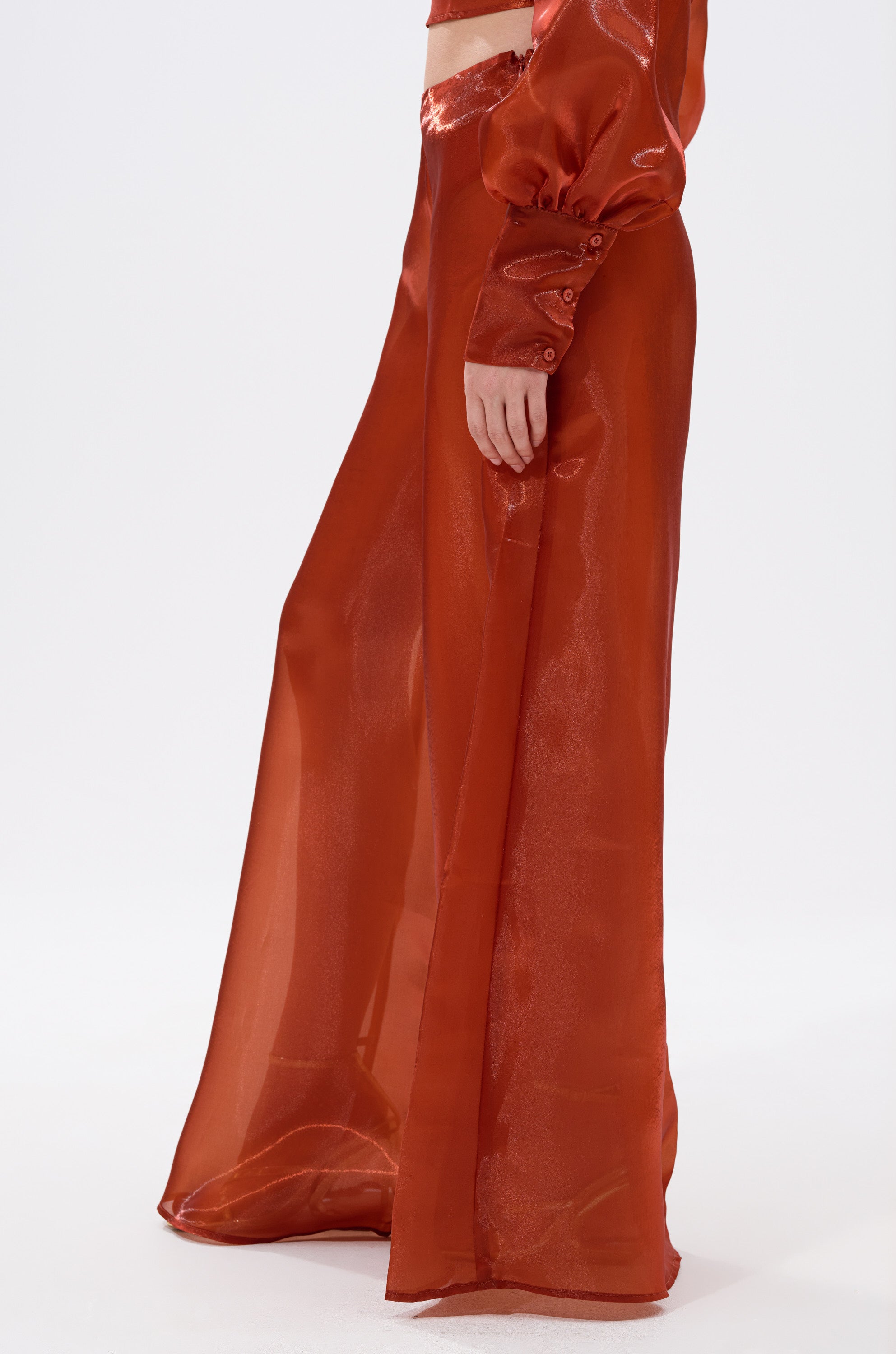 A person wears the FEELING FREE CHIFFON PALAZZO PANT in sheer, flowing rust-orange chiffon with a wide-leg fit, paired with a matching long-sleeve top. The shiny, translucent fabric stands out against the plain white background. 1