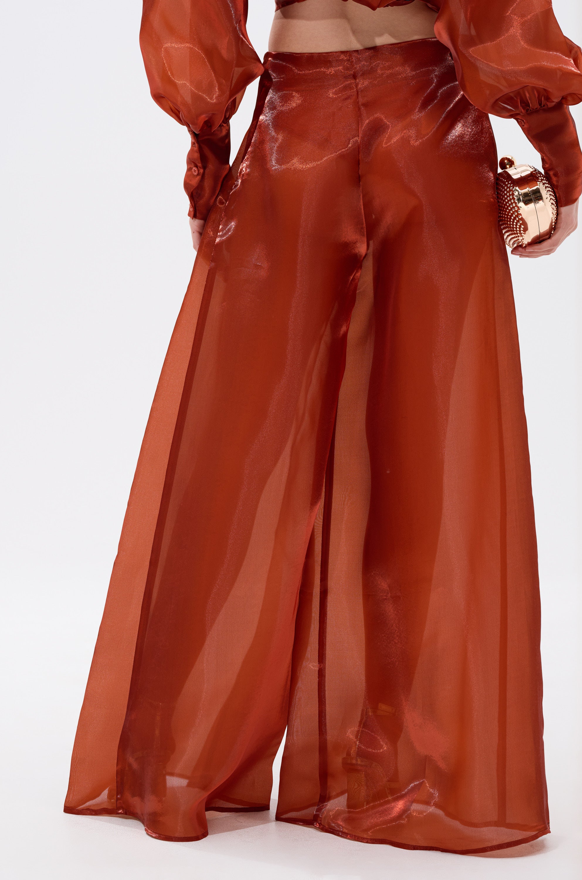 A person wears the FEELING FREE CHIFFON PALAZZO PANT in sheer, shiny rust chiffon with a matching top, holding a small metallic clutch; the image highlights their lower back and legs. 2