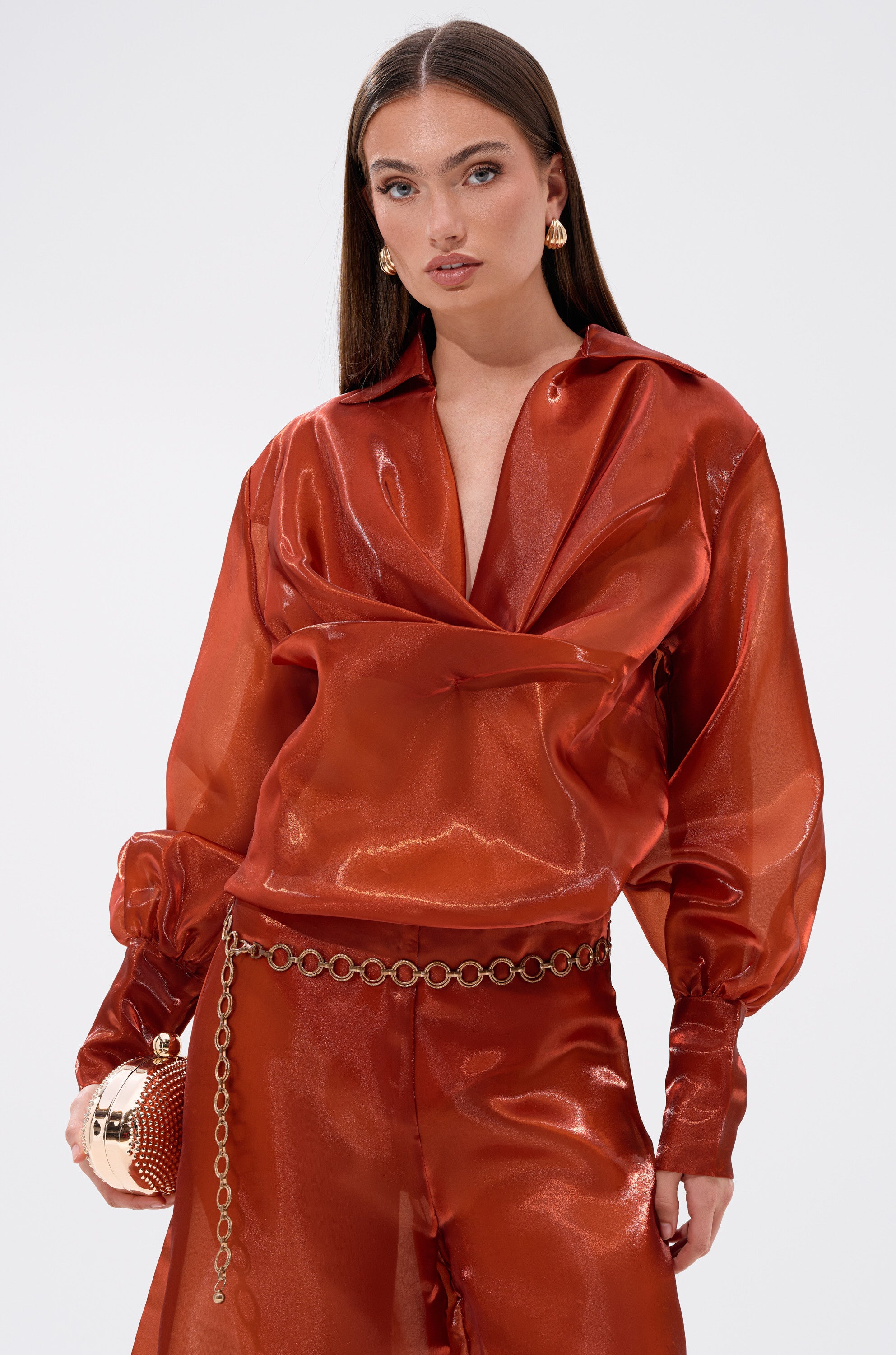 A woman with long brown hair wears the FEELING FREE CHIFFON BLOUSE in deep red with matching pants, styled with gold hoop earrings, a chain belt, and a gold bag, posing against a plain light background.
