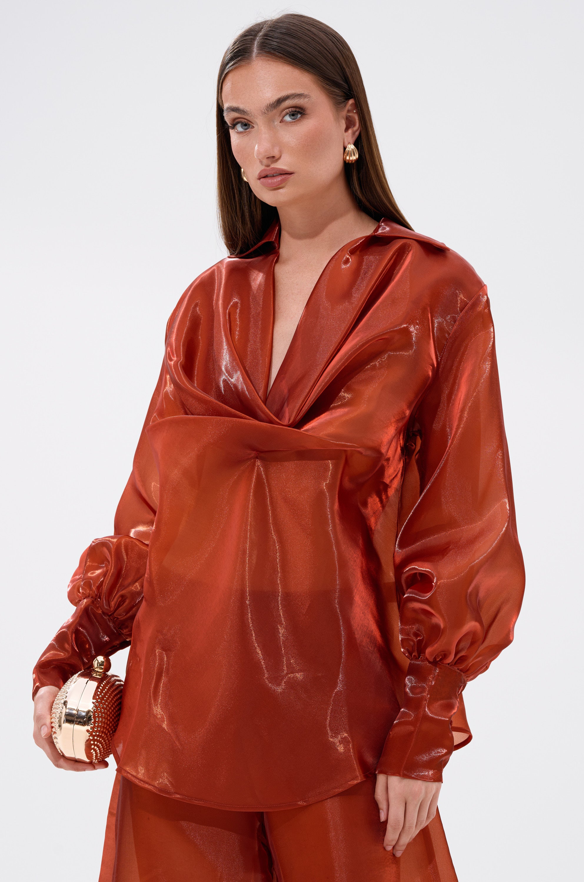 A woman with straight brown hair wears the FEELING FREE CHIFFON BLOUSE in shiny, sheer burnt-orange. With voluminous sleeves and matching pants, she confidently holds a small metallic clutch against a plain background.