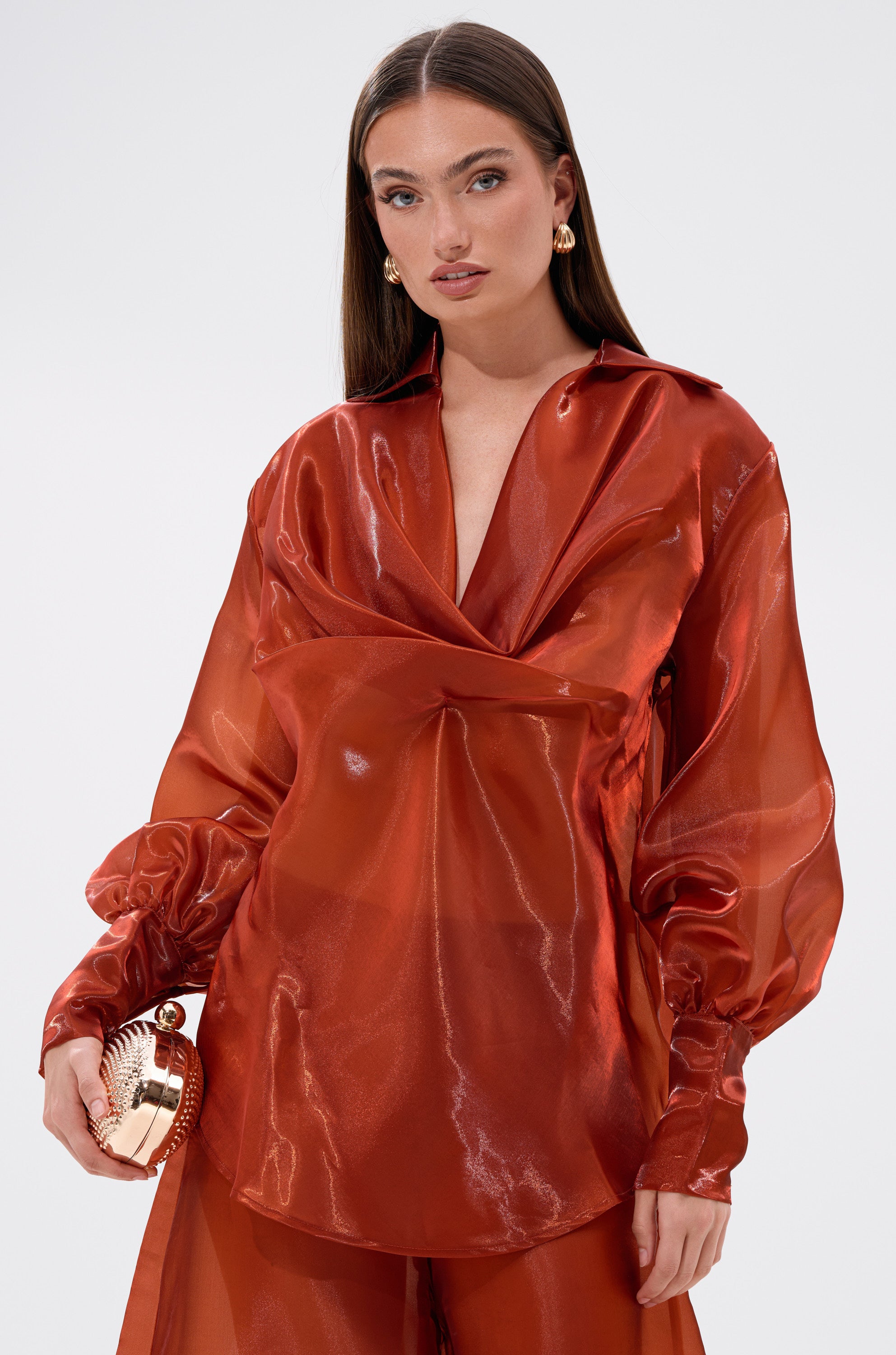 A woman with long brown hair wears the FEELING FREE CHIFFON BLOUSE in shiny, sheer red with a deep neckline and matching pants. She holds a metallic clutch and stands against a plain light background.