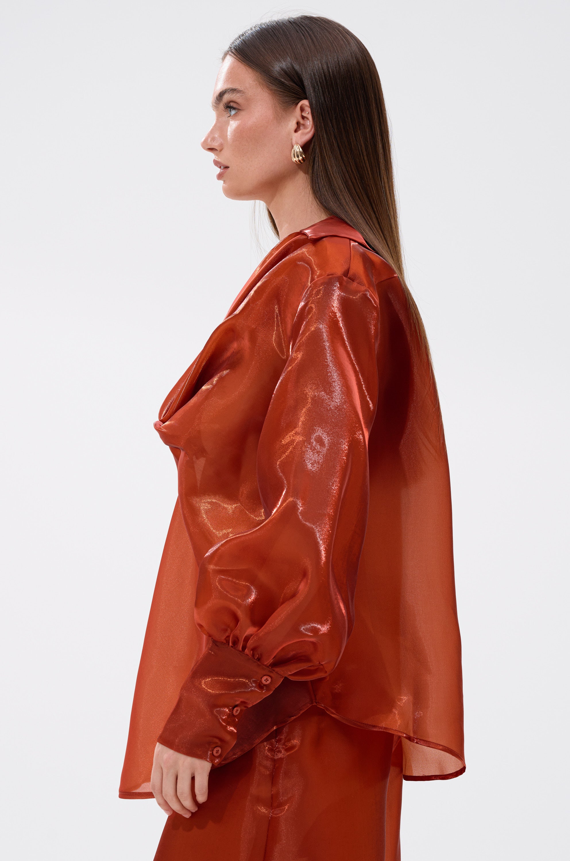 A woman with straight brown hair is shown in profile wearing the FEELING FREE CHIFFON BLOUSE—a shiny, sheer, rust chiffon top with long puffed sleeves and matching pants—against a plain light background.