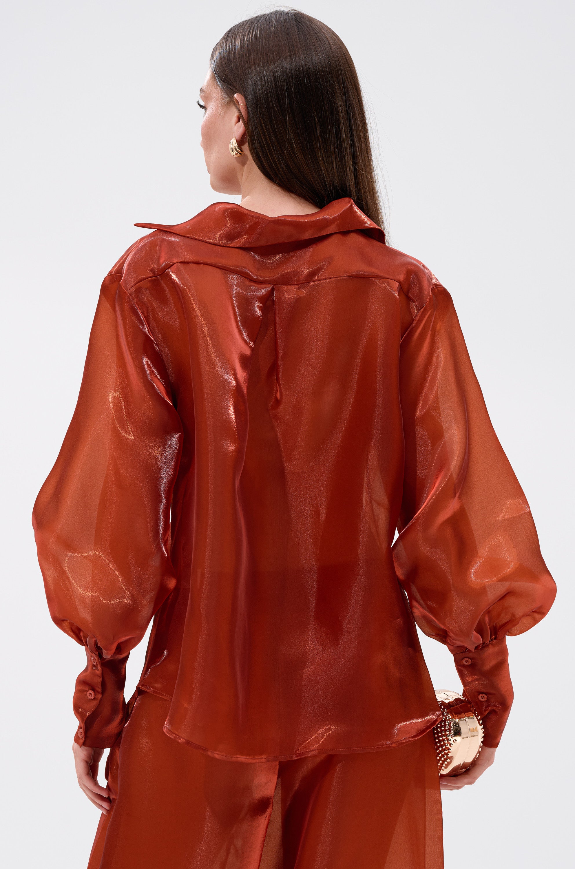 A woman with long brown hair, seen from behind, wears the FEELING FREE CHIFFON BLOUSE in a shiny, sheer rust shade with puffy sleeves and matching pants, holding a small round gold clutch in her left hand.