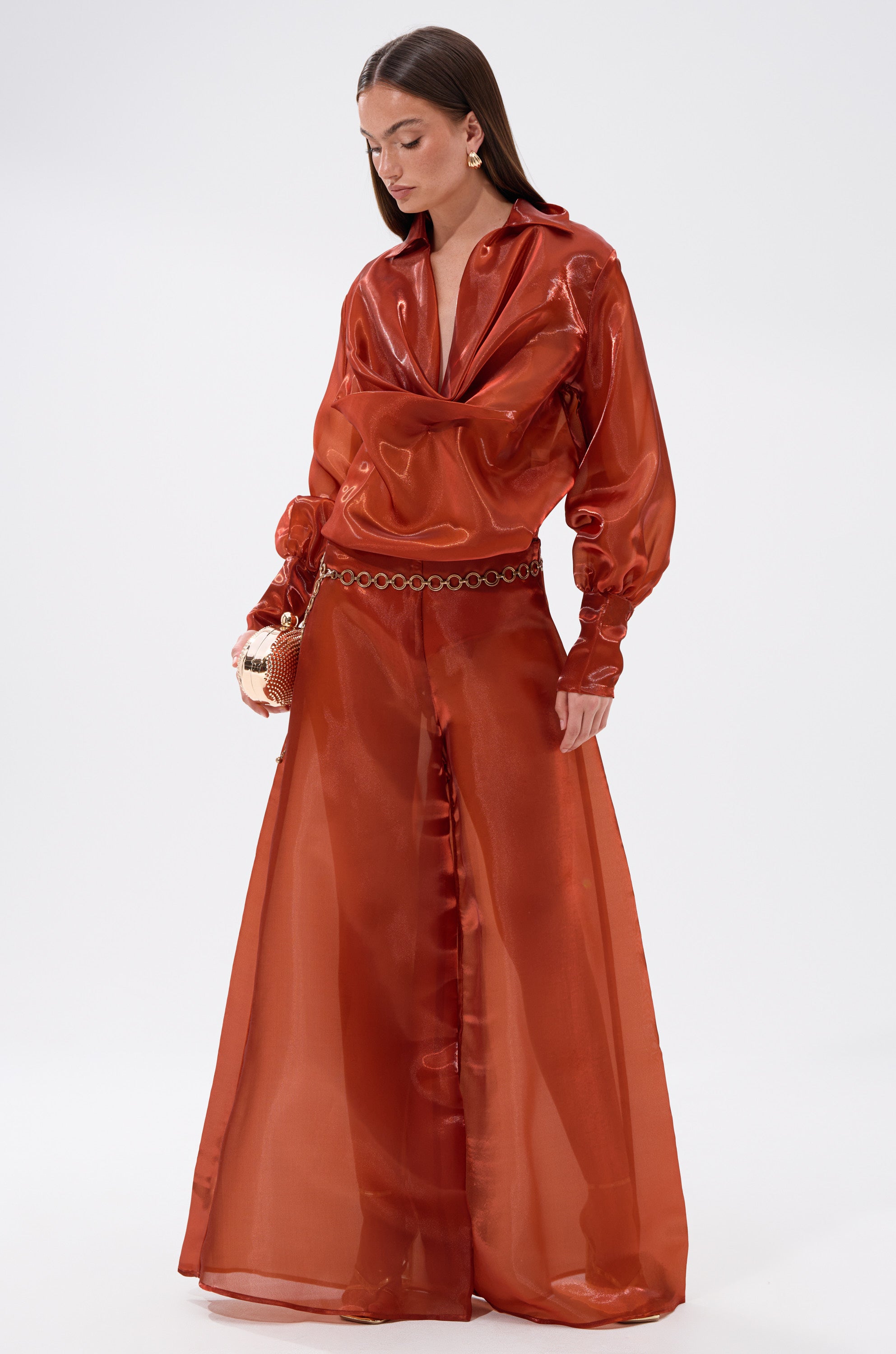 A woman poses confidently against a white background wearing the FEELING FREE CHIFFON BLOUSE in shiny, sheer burnt orange with wide-leg pants, statement earrings, and a small handbag.