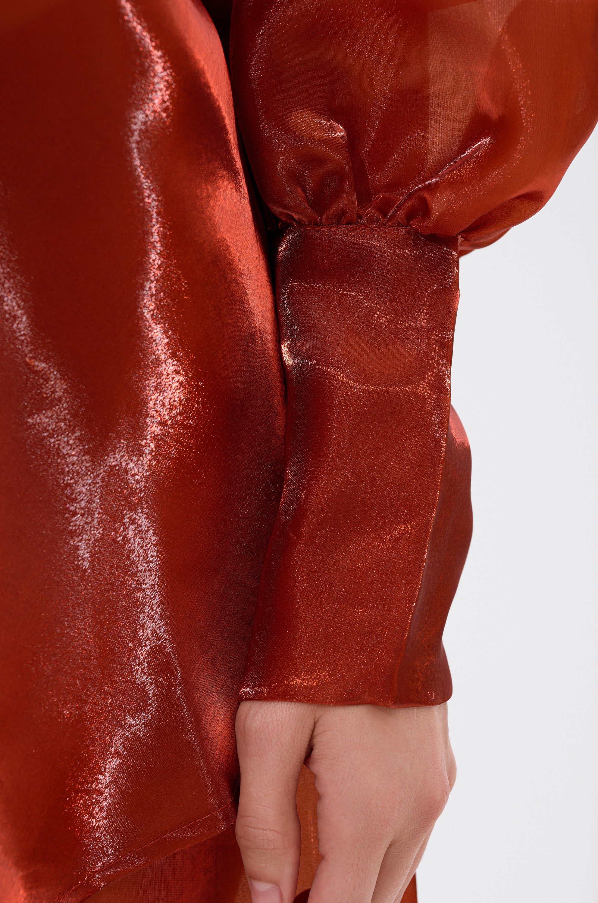 A close-up of a hand resting against the body, wearing the shiny, metallic red FEELING FREE CHIFFON BLOUSE with gathered cuffs. The fabric catches light, creating brilliant highlights.