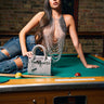 A woman in the SHAKE IT FOR ME BABY BEADED BLOUSE and ripped jeans sits on a pool table, with a silver jeweled handbag nearby against an exposed brick wall.