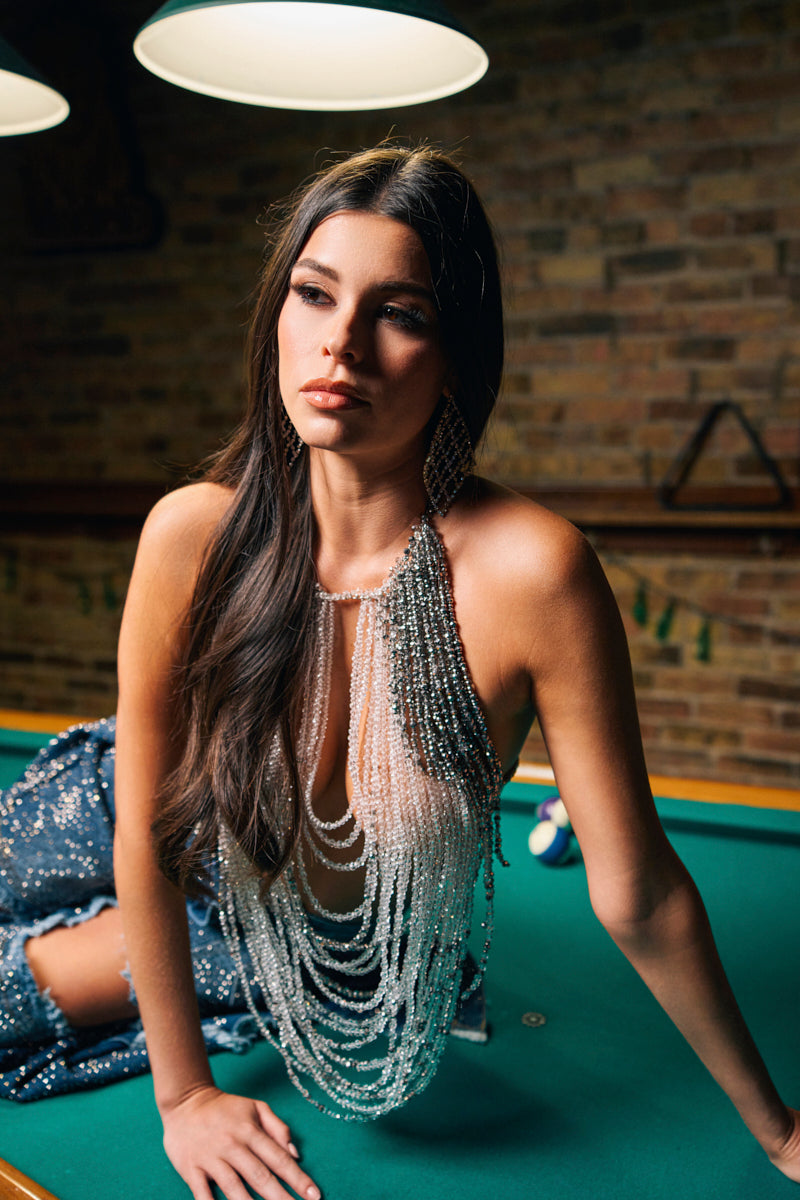 A woman with long dark hair wears the SHAKE IT FOR ME BABY BEADED BLOUSE while sitting on a pool table under hanging lights, gazing thoughtfully to the side. Brick walls and billiard equipment decorate the background.