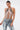 A woman poses confidently in the SHAKE IT FOR ME BABY BEADED BLOUSE featuring silver and white glass beading, styled with light blue lace-up jeans, slicked-back hair, and dangling earrings against a plain white background.
