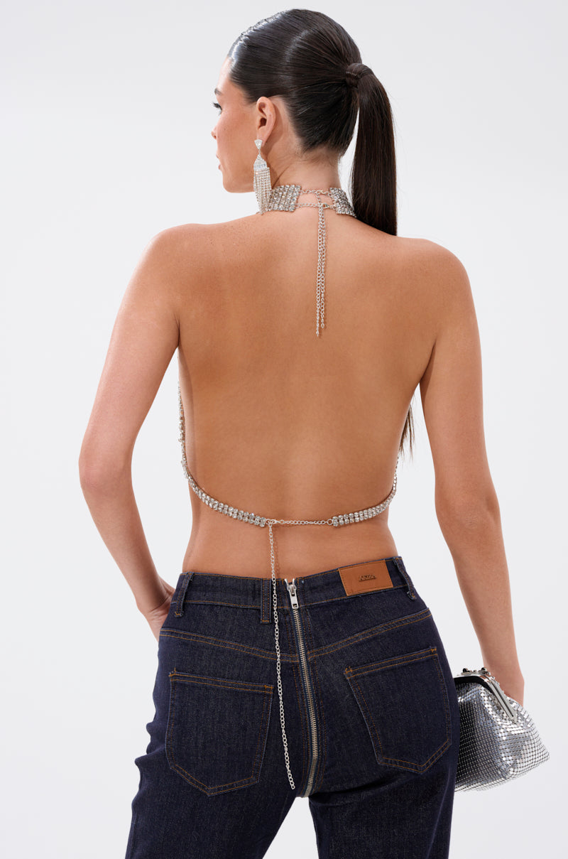 A woman with a sleek ponytail stands with her back to the camera, wearing the ALL EYES ON ME HALTER BLOUSE with sparkling straps, dark blue jeans, and holding a silver mesh handbag.