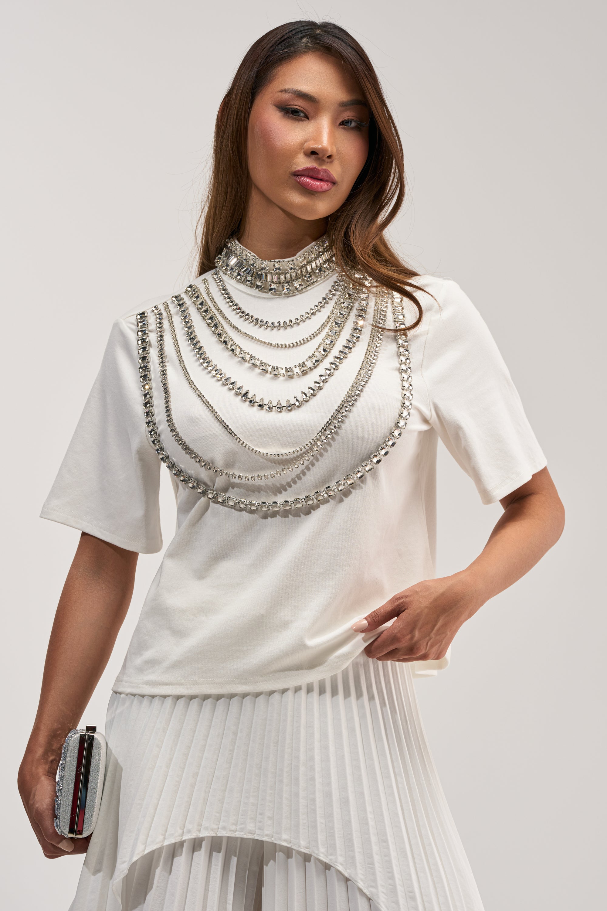 A woman in a white pleated skirt and the ROYALTY RHINESTONE TEE IN WHITE, adorned with layered silver chains and holding a small clutch, stands confidently against a plain light background.