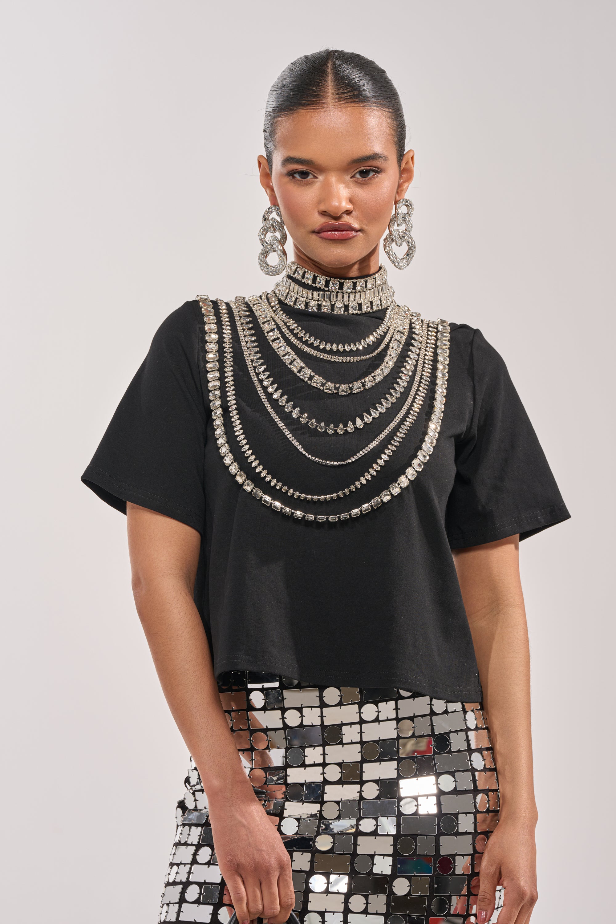 A woman stands against a light background, wearing the ROYALTY RHINESTONE TEE with layered silver statement necklaces, bold earrings, and a shiny skirt adorned with silver square mirrors. Her hair is neatly slicked back. 0