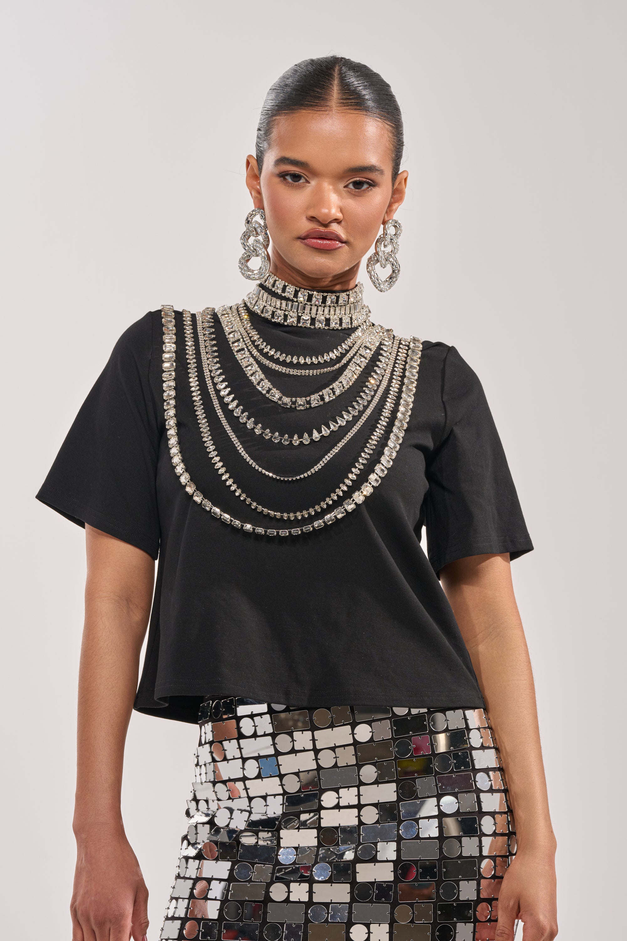 A woman wearing the ROYALTY RHINESTONE TEE, paired with layered silver necklaces, statement earrings, and a mirrored skirt, stands against a plain light background. 1