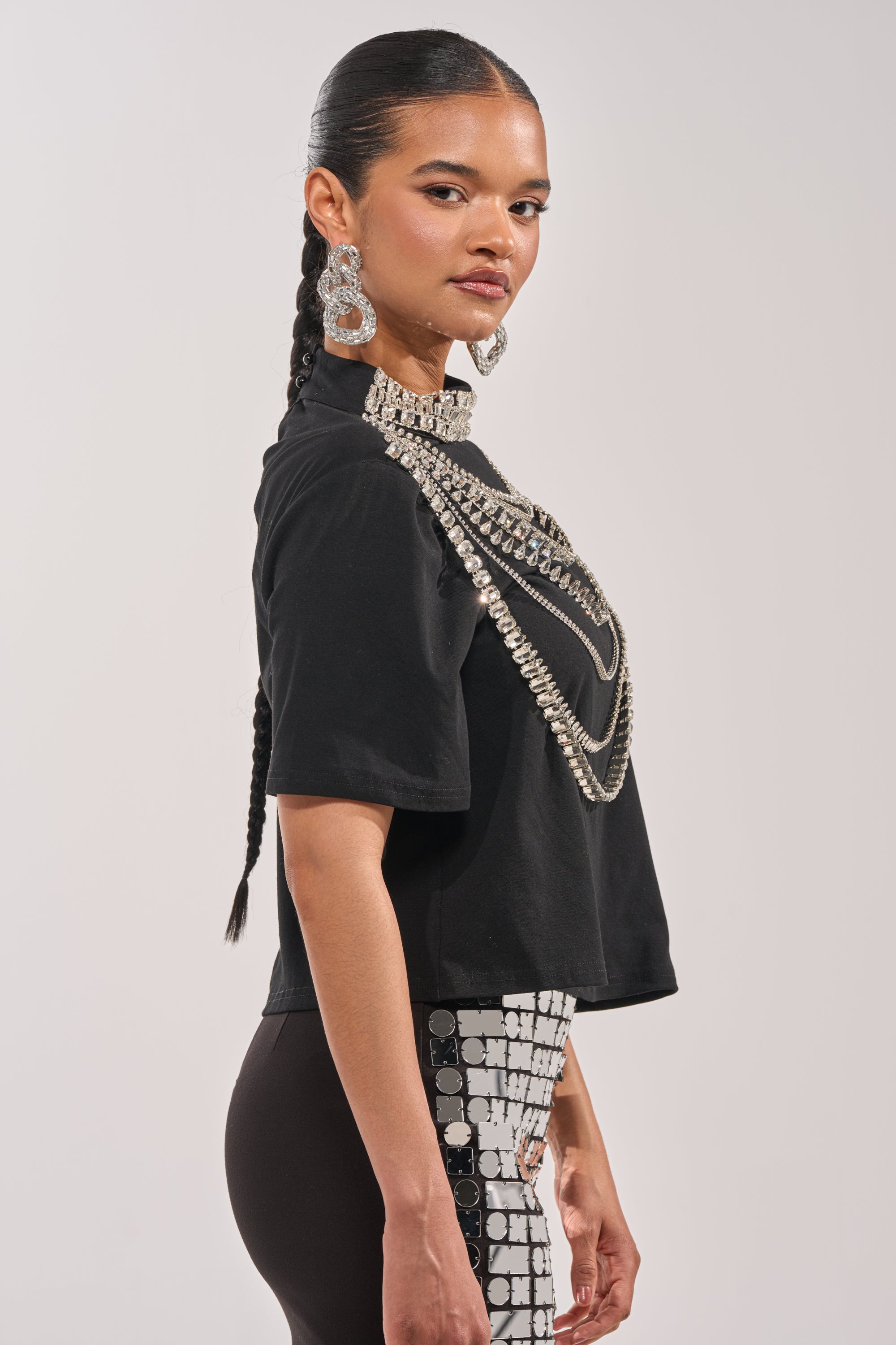 Wearing the ROYALTY RHINESTONE TEE, a woman with long dark braided hair pairs it with sparkling earrings and black pants featuring a silver square sequin pattern on one side, posing in profile. 2