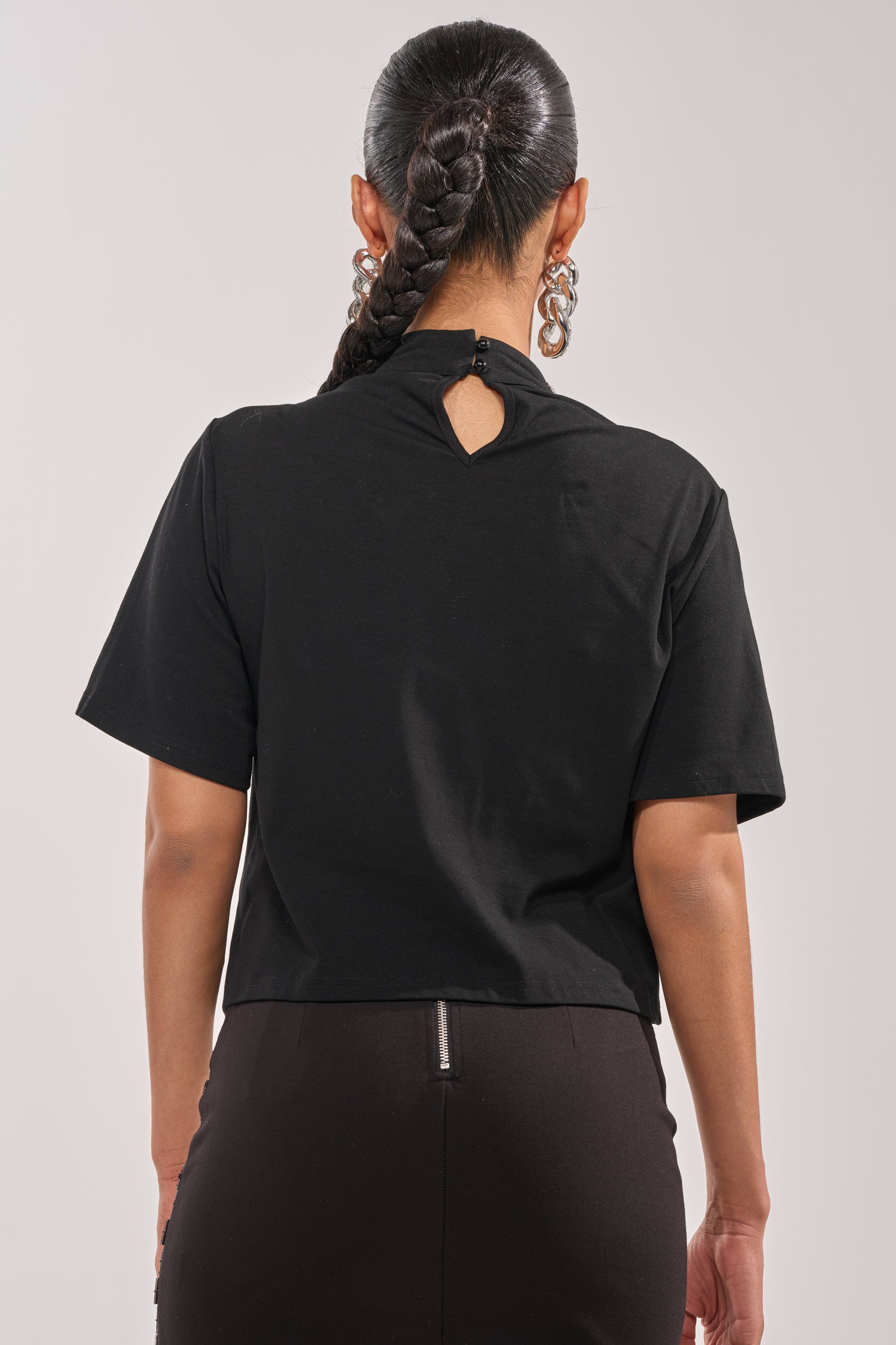 A person with long, braided hair faces away, wearing the ROYALTY RHINESTONE TEE featuring a buttoned keyhole neck and a black skirt with a visible zipper, accessorized with large hoop earrings. 3