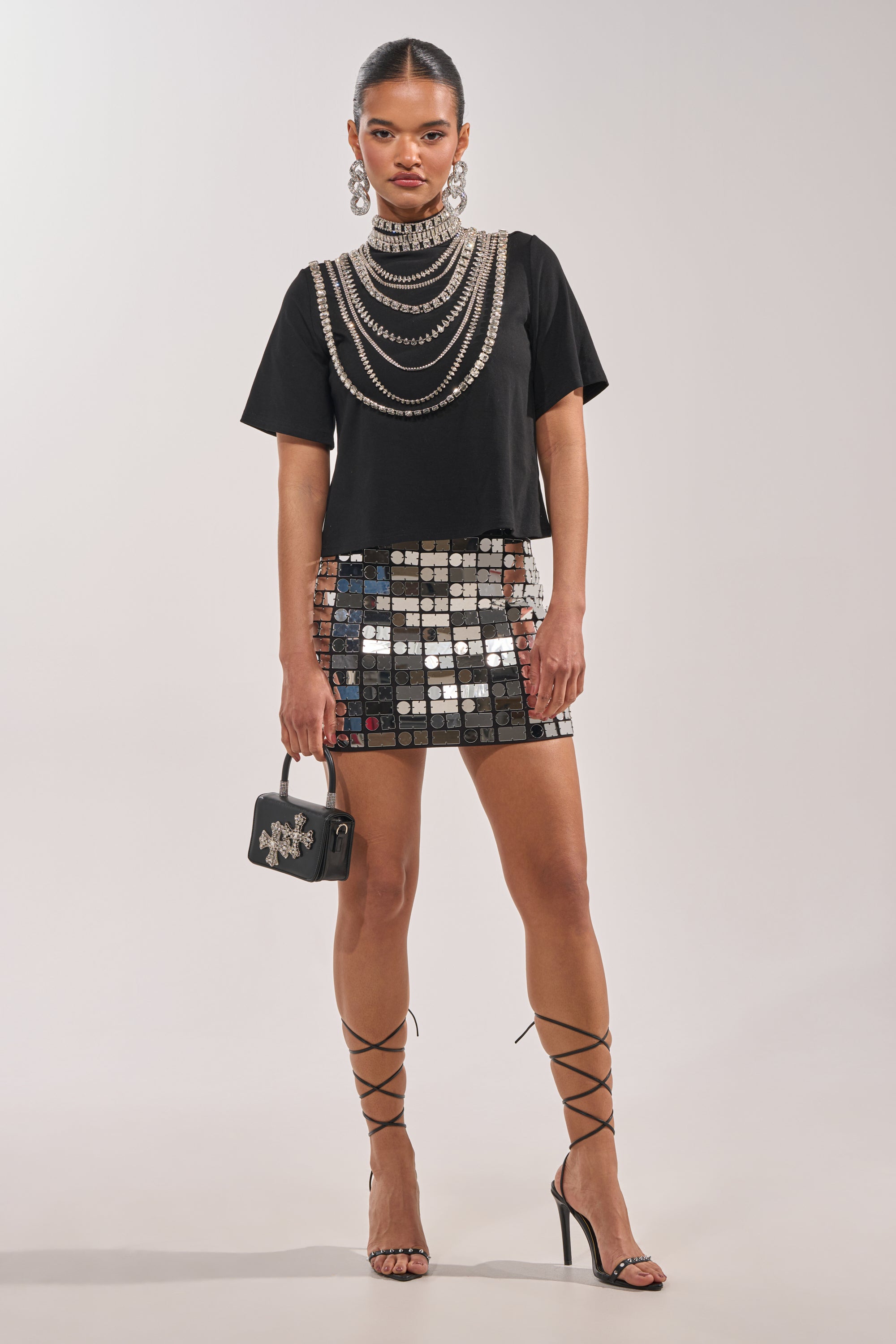 A woman poses in a studio wearing the ROYALTY RHINESTONE TEE, layered pearl necklaces, a mirrored mini skirt, lace-up heels, large earrings, and holding a small black handbag. 4