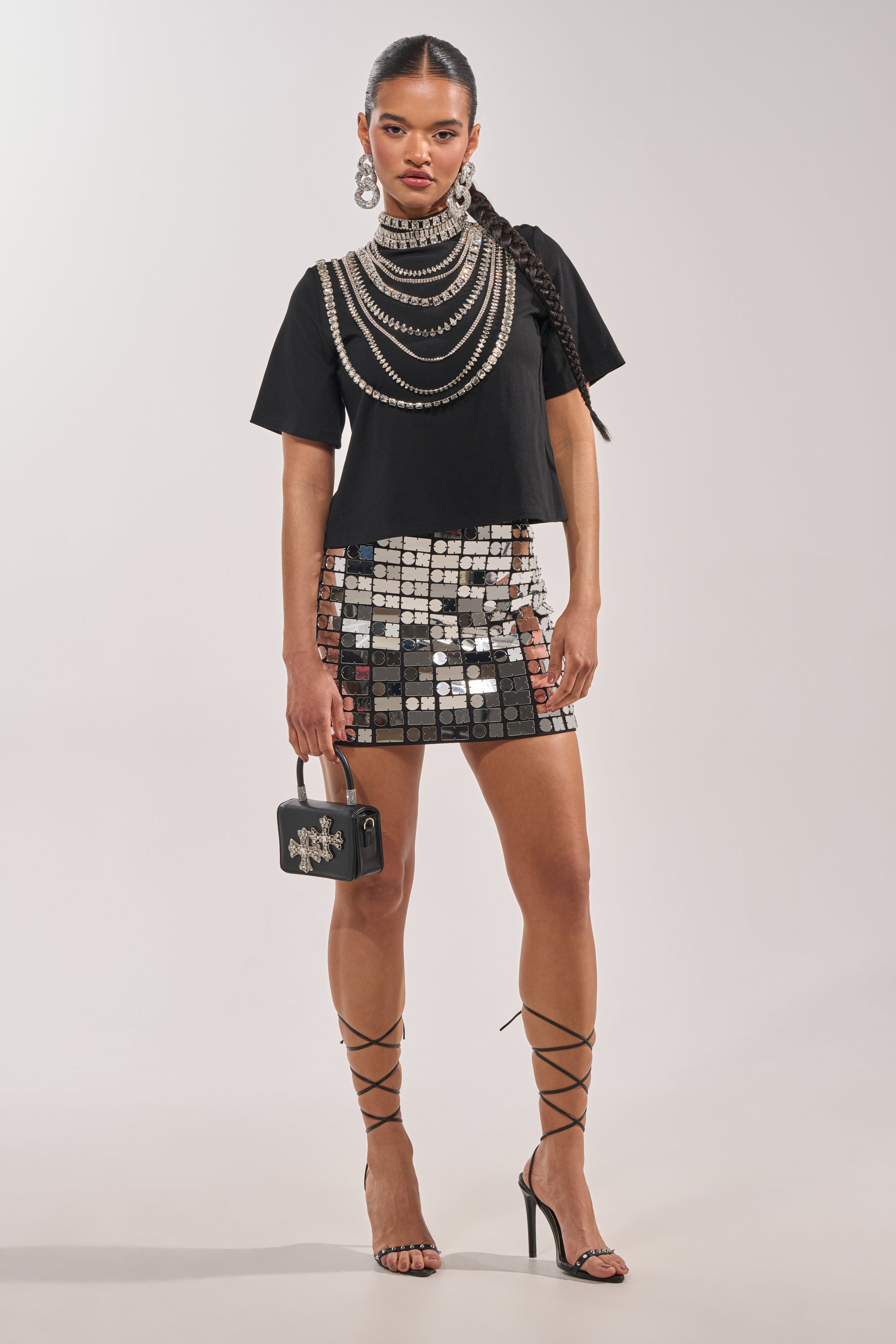 A woman in high heels wears the ROYALTY RHINESTONE TEE with layered silver necklaces, a mirror-tile mini skirt, a small black handbag, long braid, and large statement earrings. 5