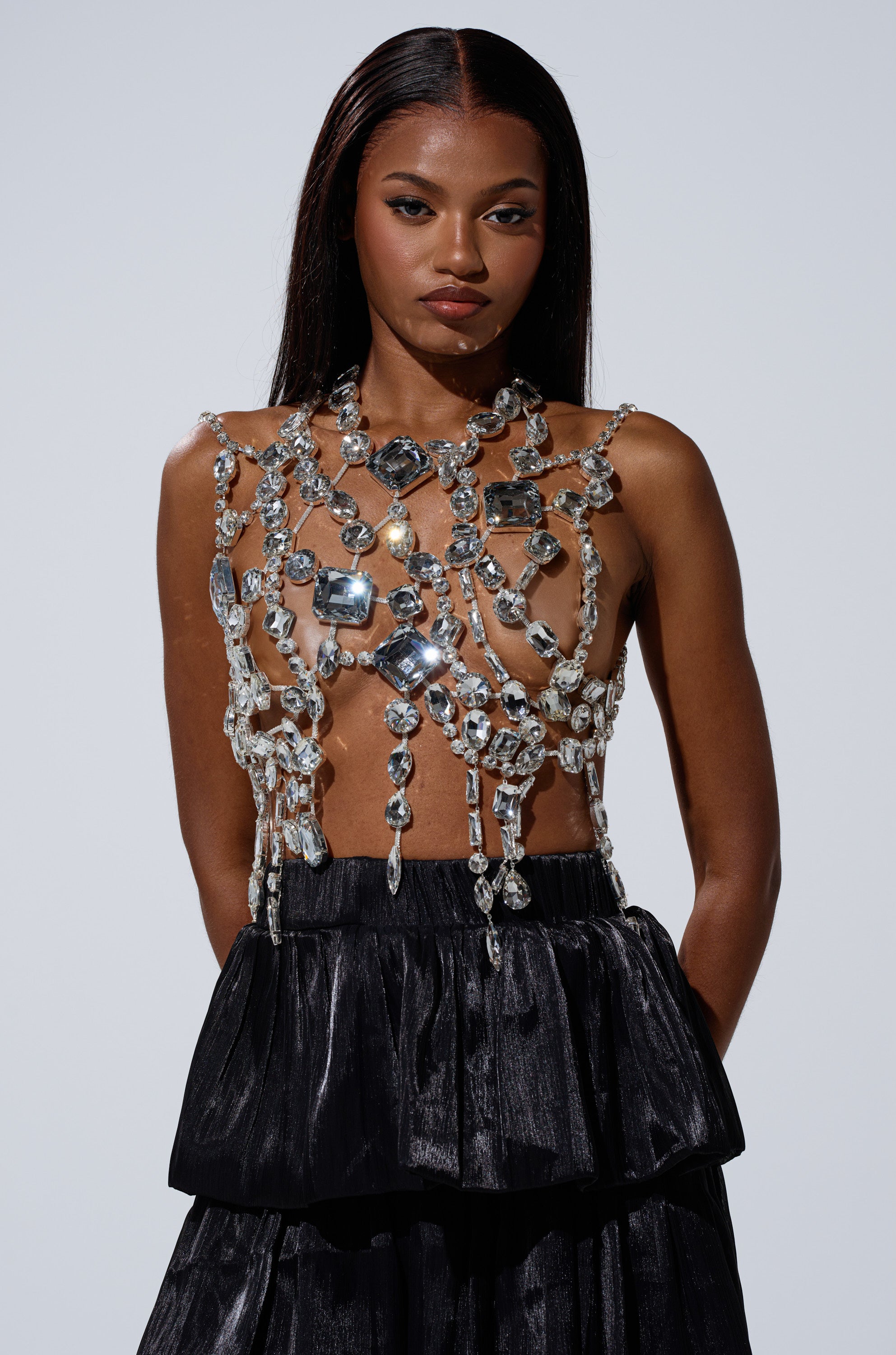 A woman with long, straight hair wears the GOTTA LET IT GO RHINESTONE BRALETTE and a black textured skirt, standing against a light gray background and looking confidently at the camera.