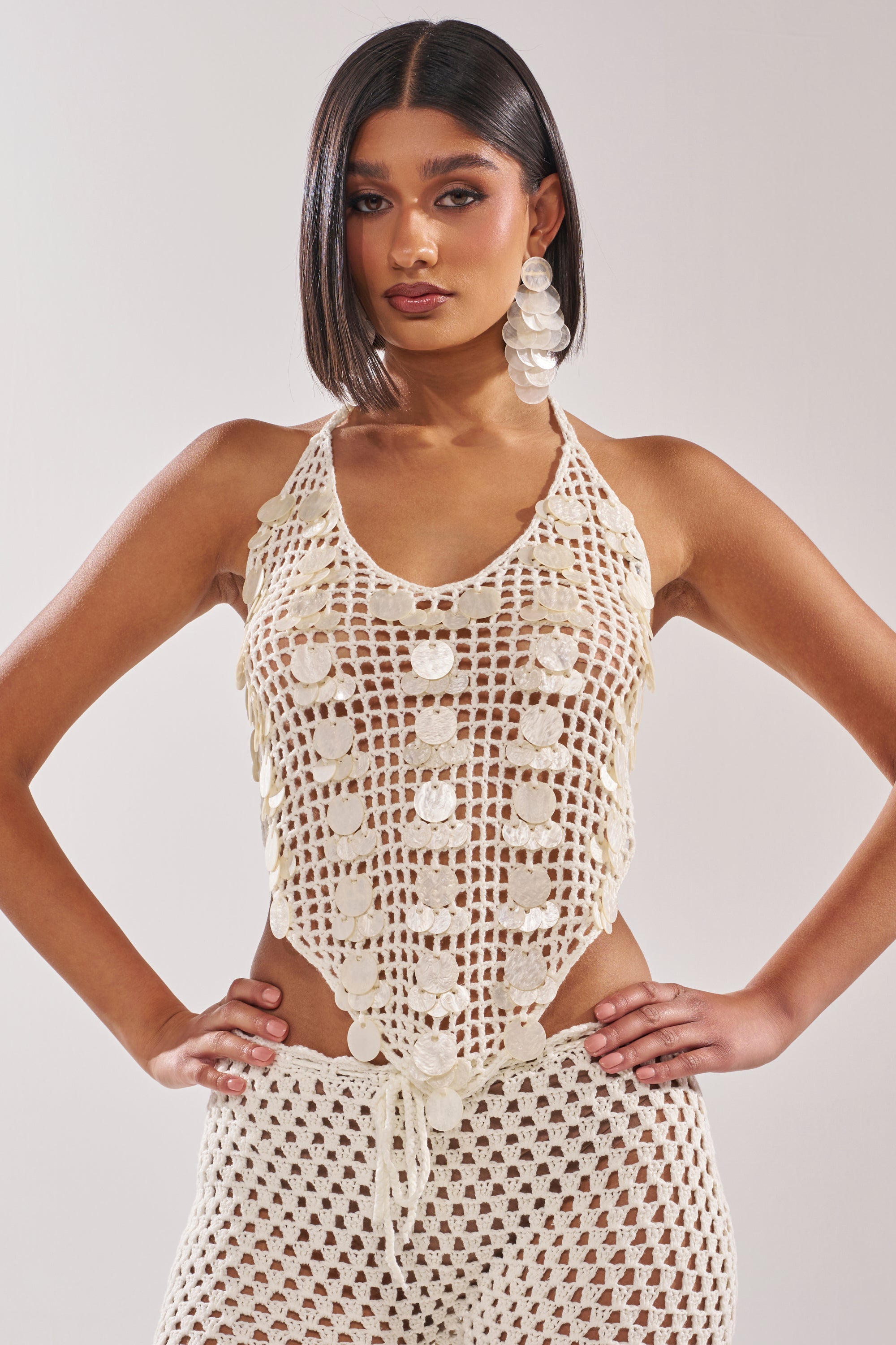 A woman with straight, dark hair wears the TAKE ME TO THE CLUB CROCHET TANK—a cream, sleeveless halter top with sequin disc accents—paired with matching crochet pants and large circular earrings, standing against a plain background. 0