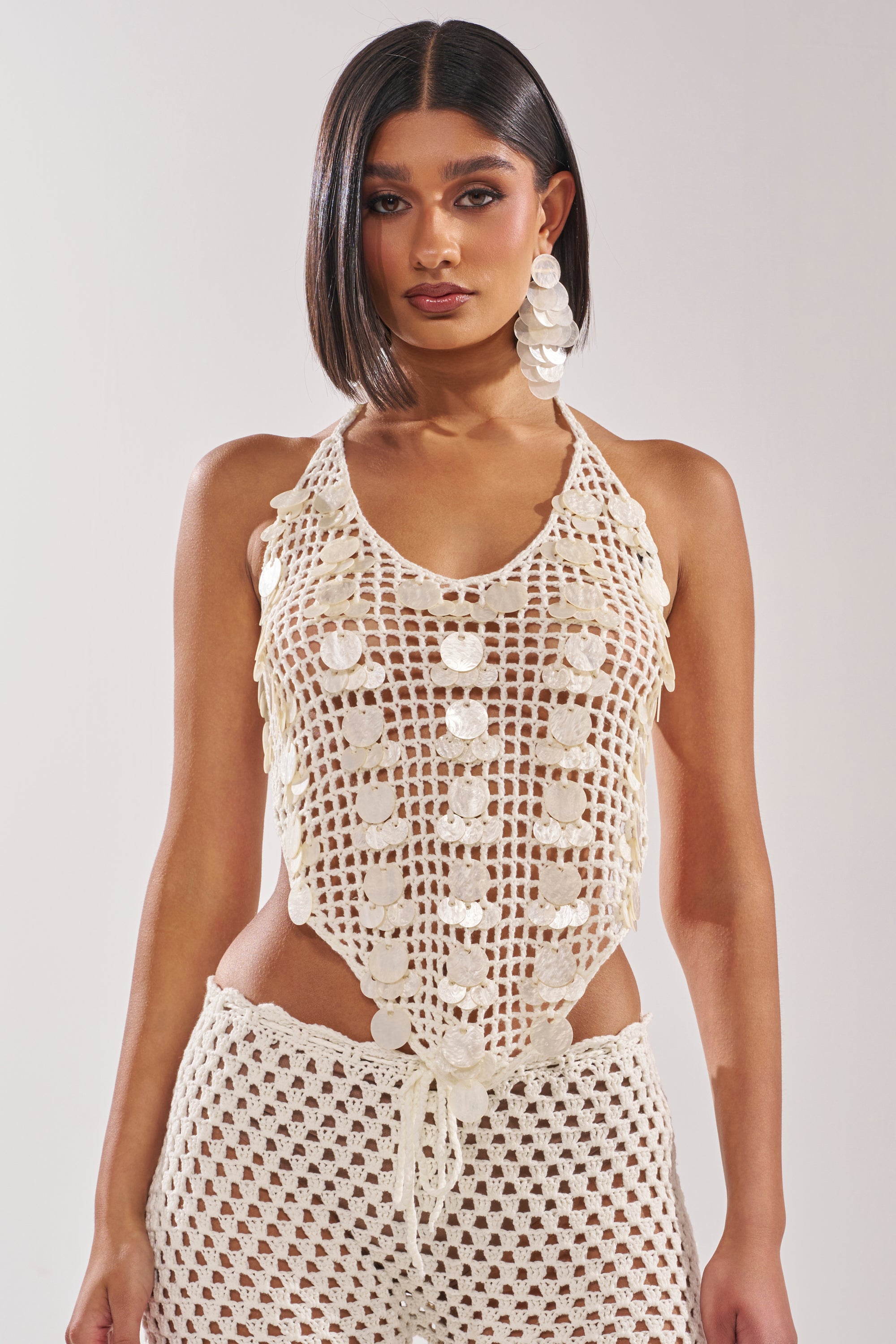 A woman with straight, shoulder-length dark hair wears the TAKE ME TO THE CLUB CROCHET TANK and matching pants with shell accents, paired with bold white earrings for a chic beach club look. 1