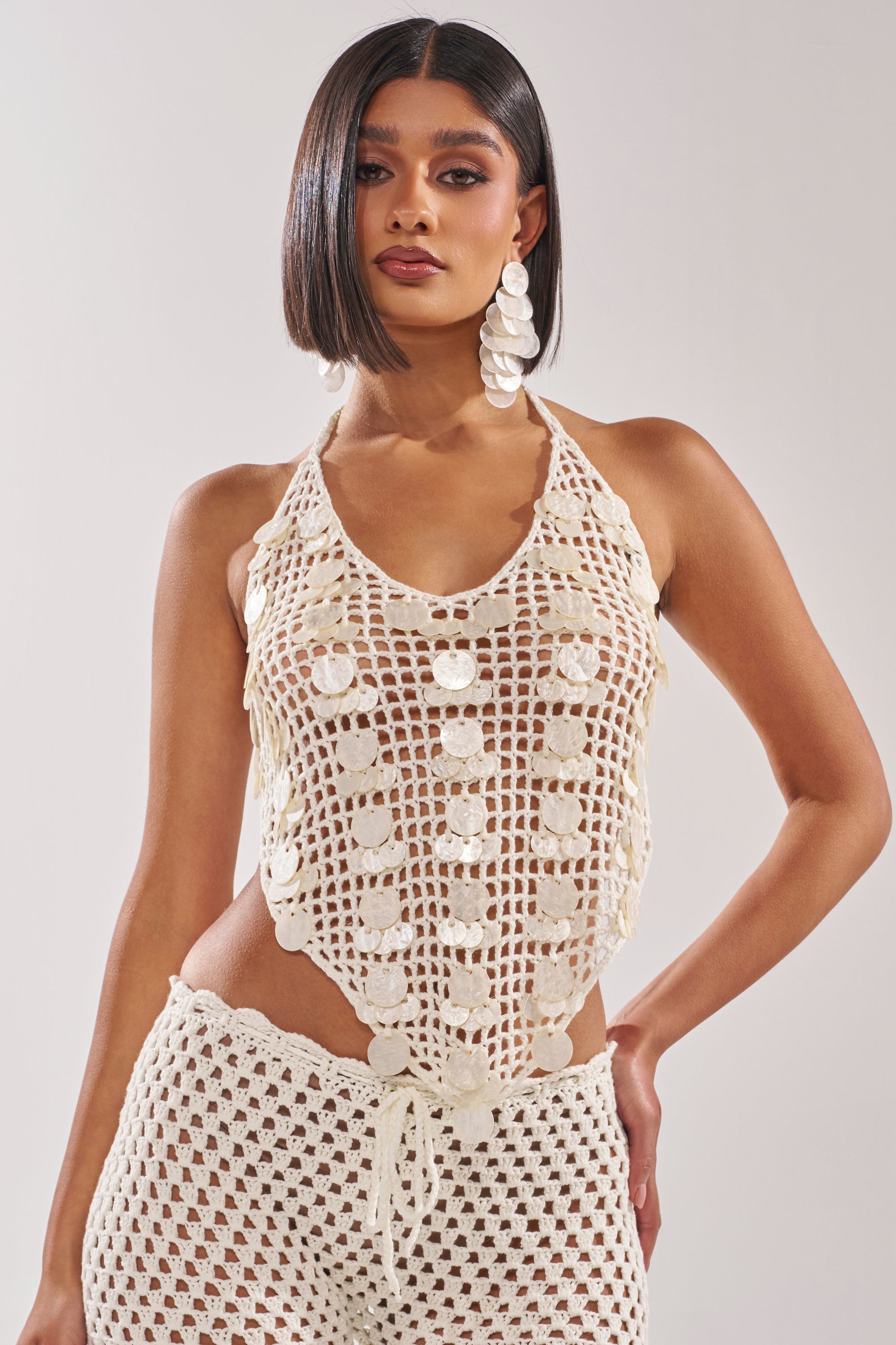 A woman with straight, shoulder-length hair wears the TAKE ME TO THE CLUB CROCHET TANK with matching pants—a chic beach club look. She accessorizes with large, round white earrings and poses against a plain light background. 2