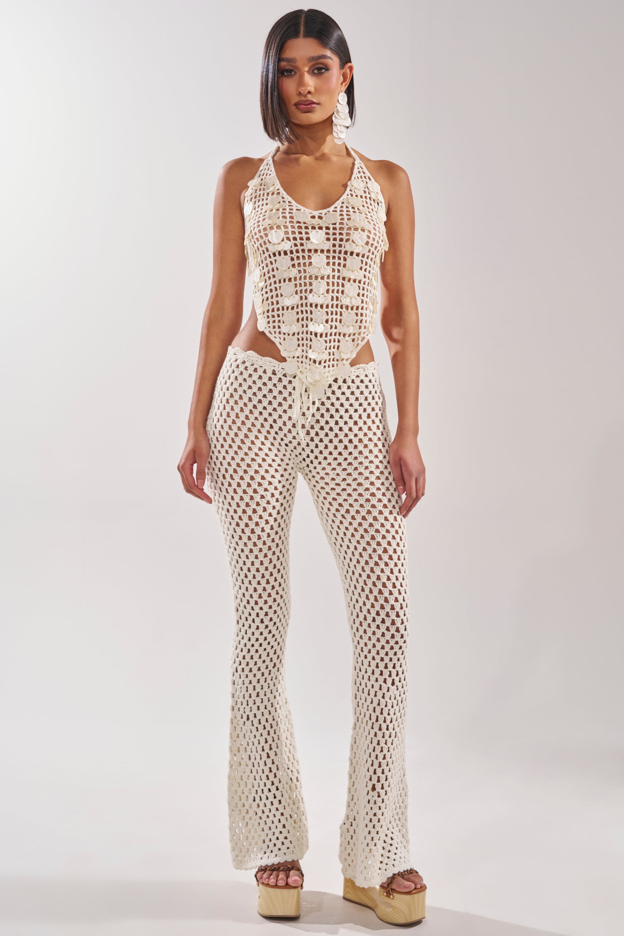 A woman faces forward in the TAKE ME TO THE CLUB CROCHET TANK, a cream-colored crocheted halter top with sequin details and matching flared pants. She has straight, shoulder-length hair and wears platform sandals against a white background. 6