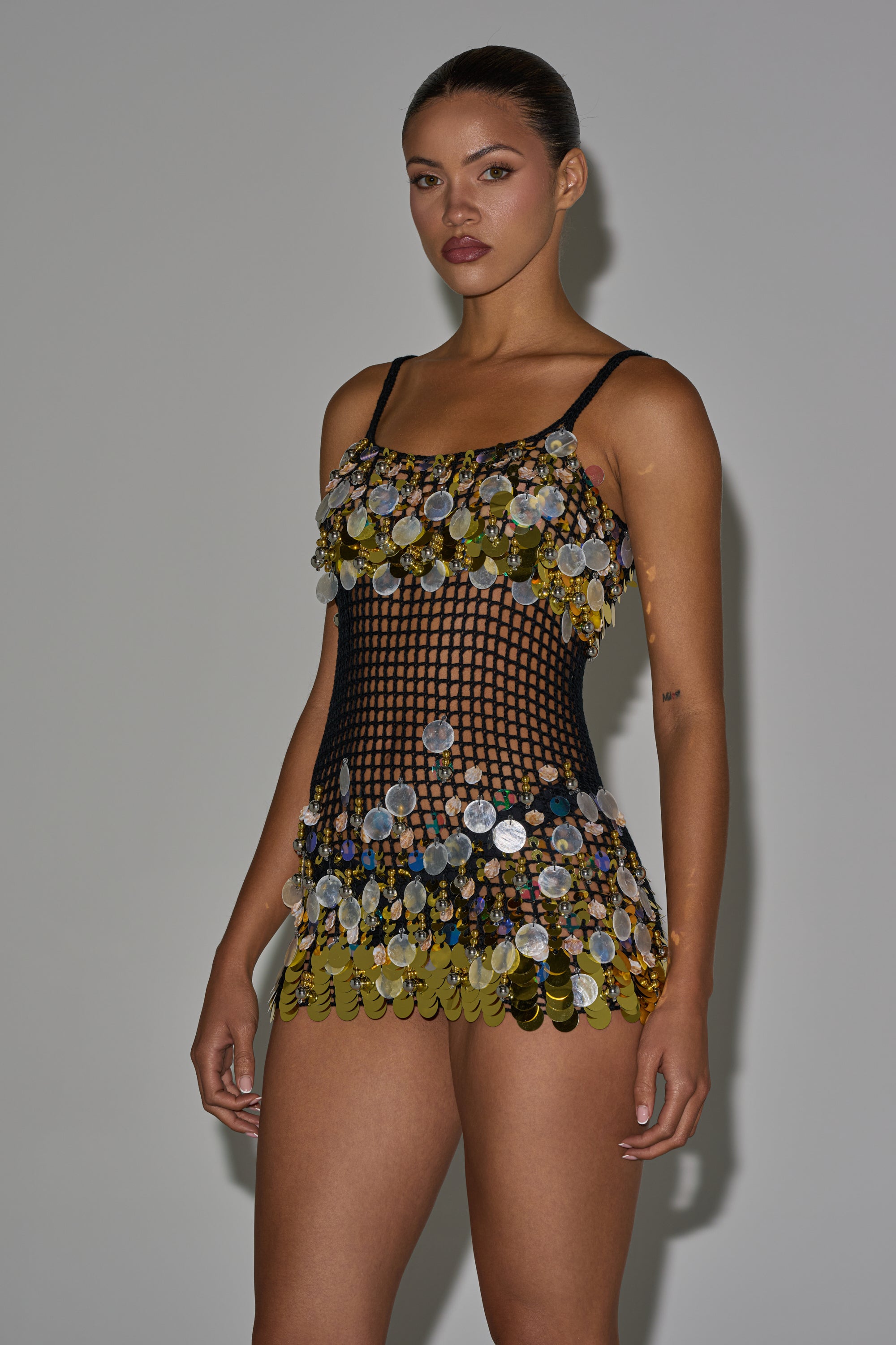A woman stands against a plain background, confidently wearing the RESORT LIVING SEQUIN CROCHET MINI DRESS—a sleeveless black mini dress embellished with bold gold and silver sequins. 0
