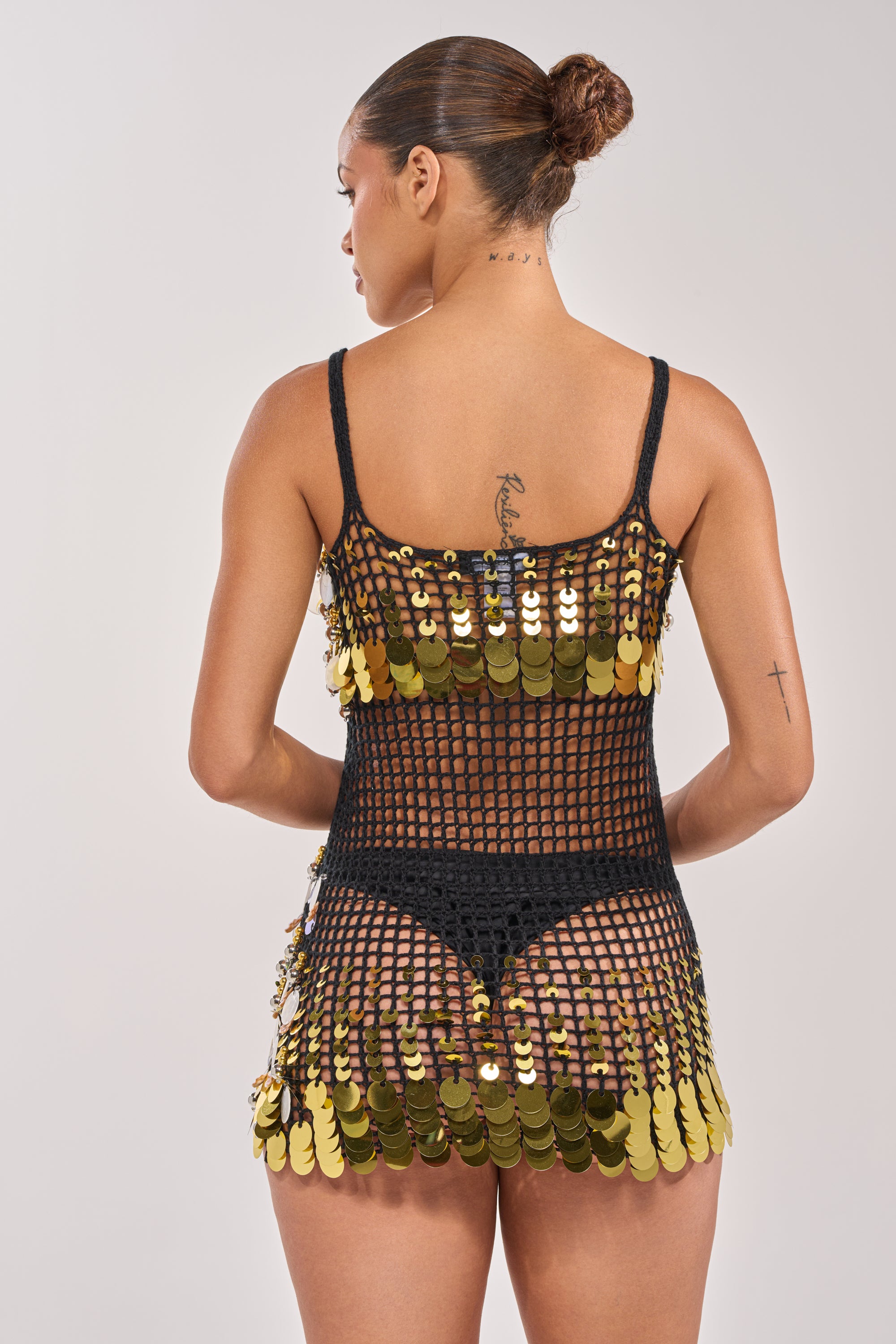 A woman with a sleek bun and visible neck and arm tattoos faces away, modeling the RESORT LIVING SEQUIN CROCHET MINI DRESS over black underwear, against a plain light background. 4
