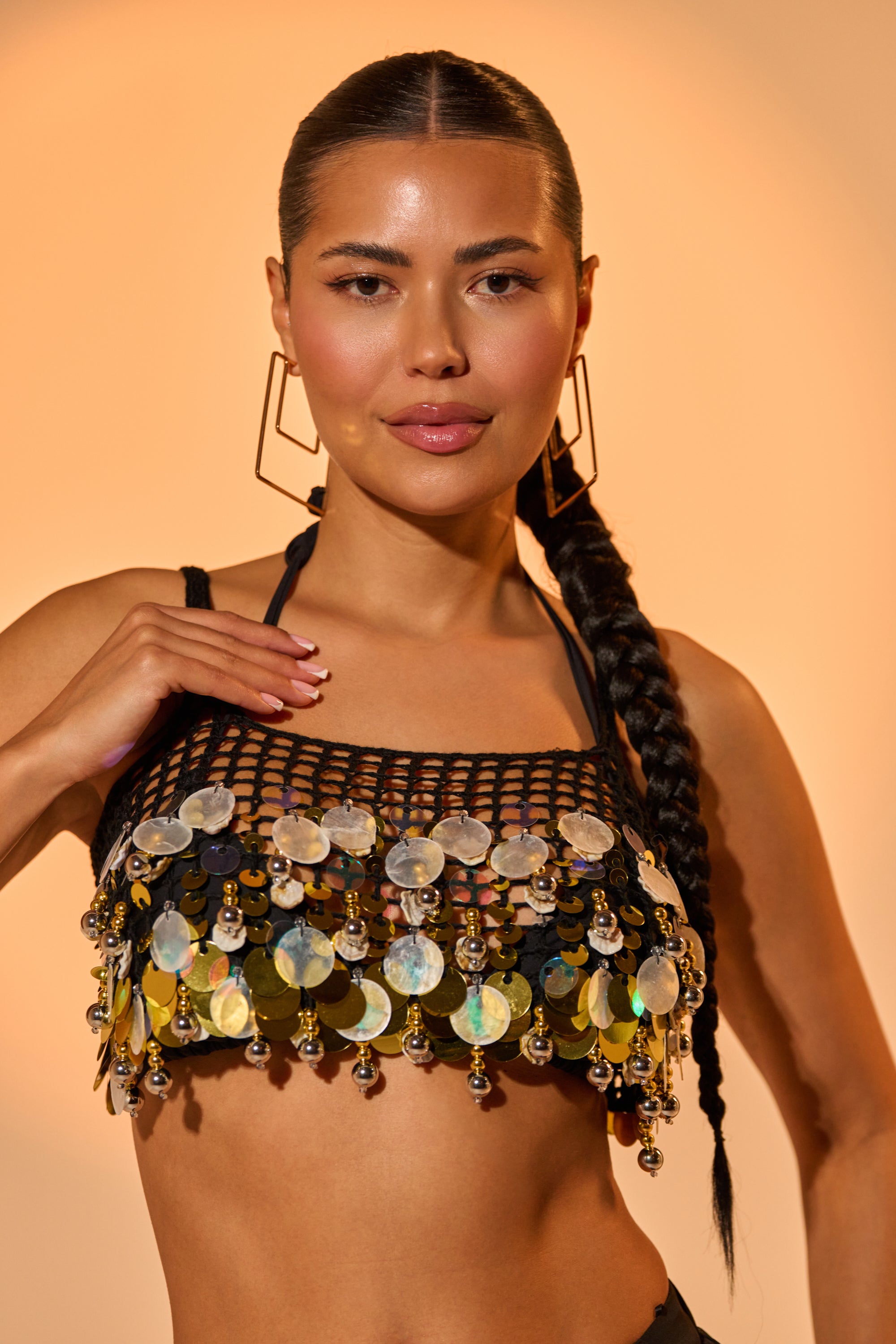 A woman with long, dark braided hair poses confidently against a warm beige background, wearing geometric earrings and the RESORT LIVING SEQUIN CROCHET TOP—a black mesh crop top with large iridescent sequins. 0