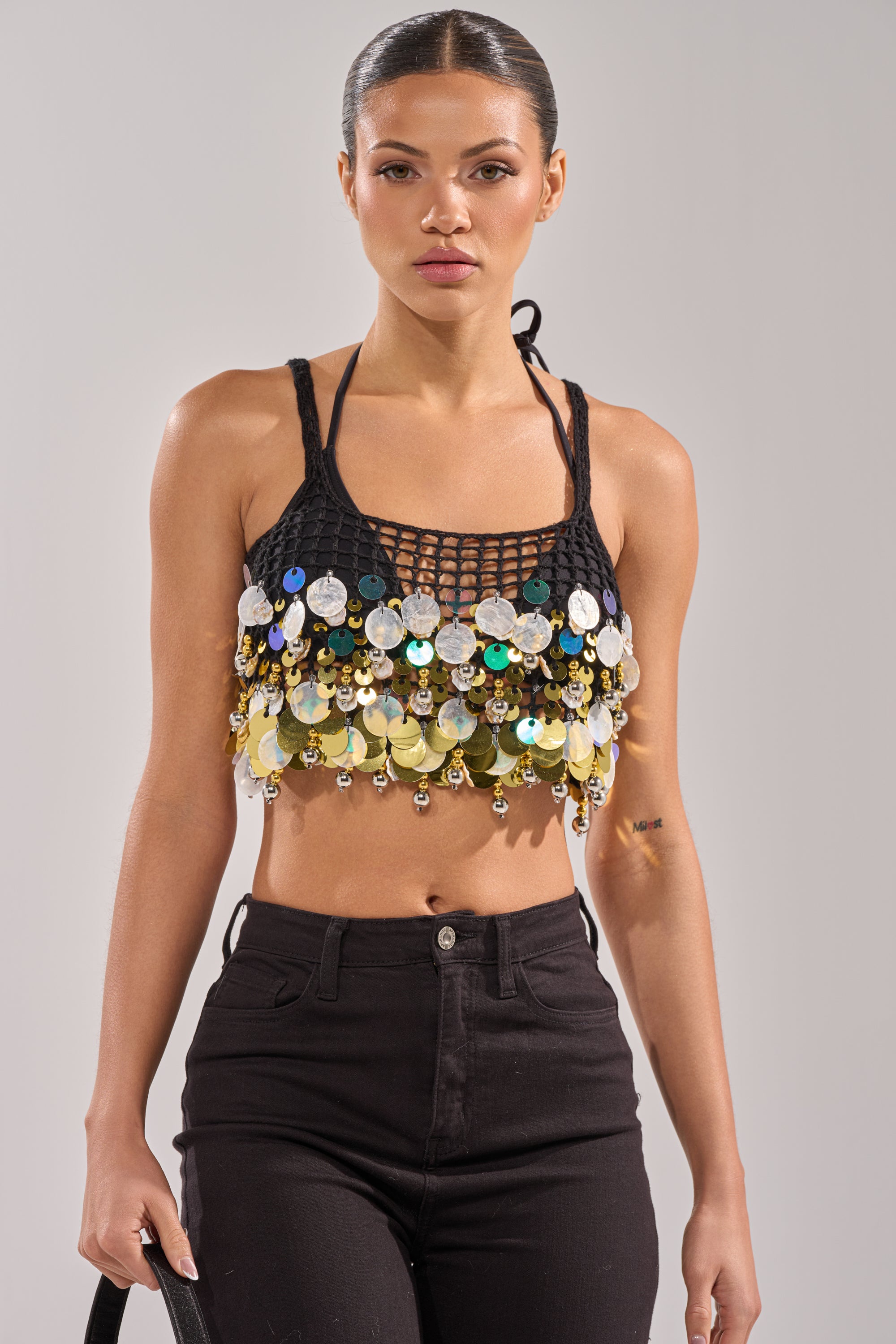 A woman wears the RESORT LIVING SEQUIN CROCHET TOP with high-waisted black jeans, posing confidently against a neutral background—an ideal choice for a festival party look. 2