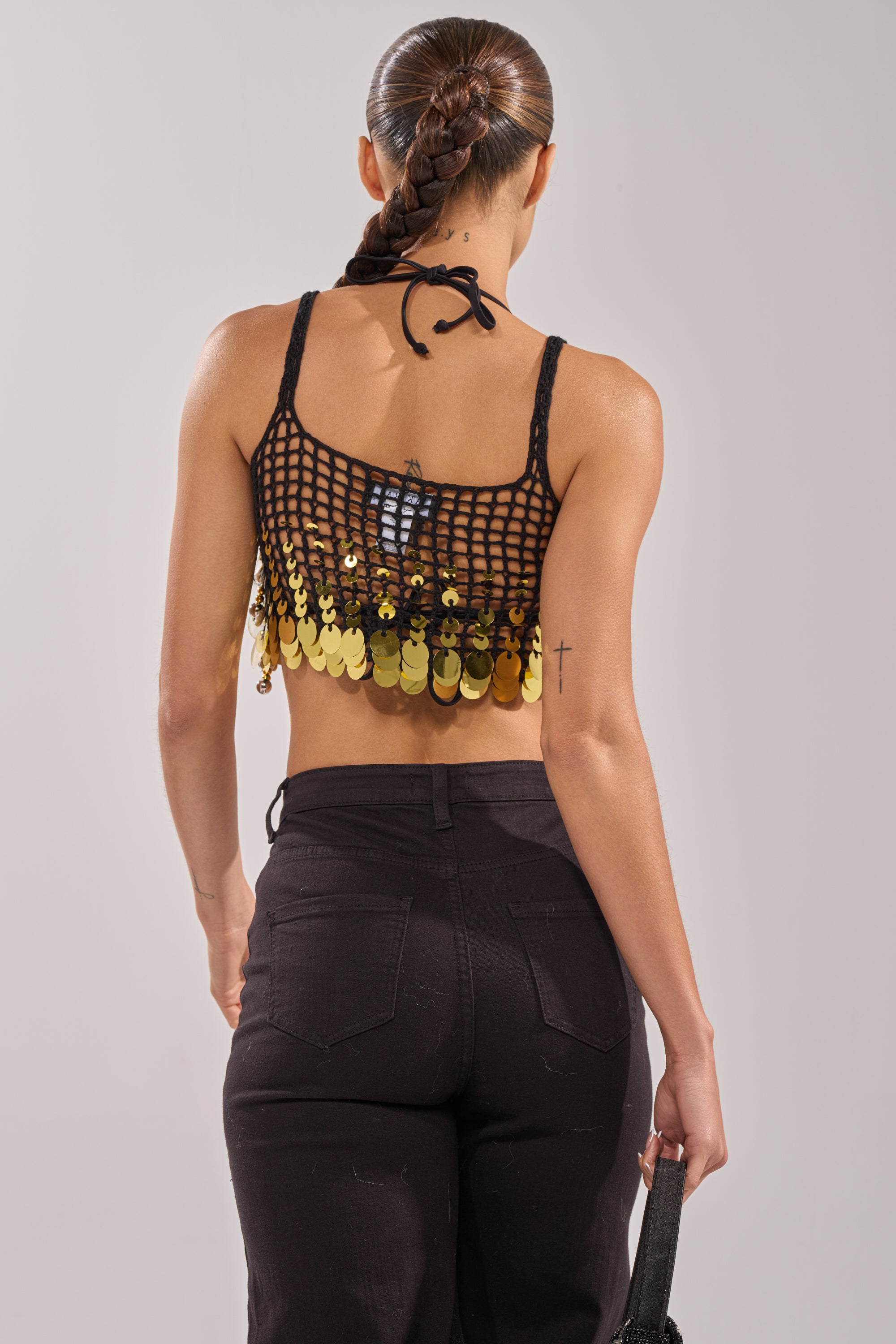A woman with a braided ponytail wears the RESORT LIVING SEQUIN CROCHET TOP and high-waisted black pants. With her back to the camera, she holds a small black bag by her side. 5