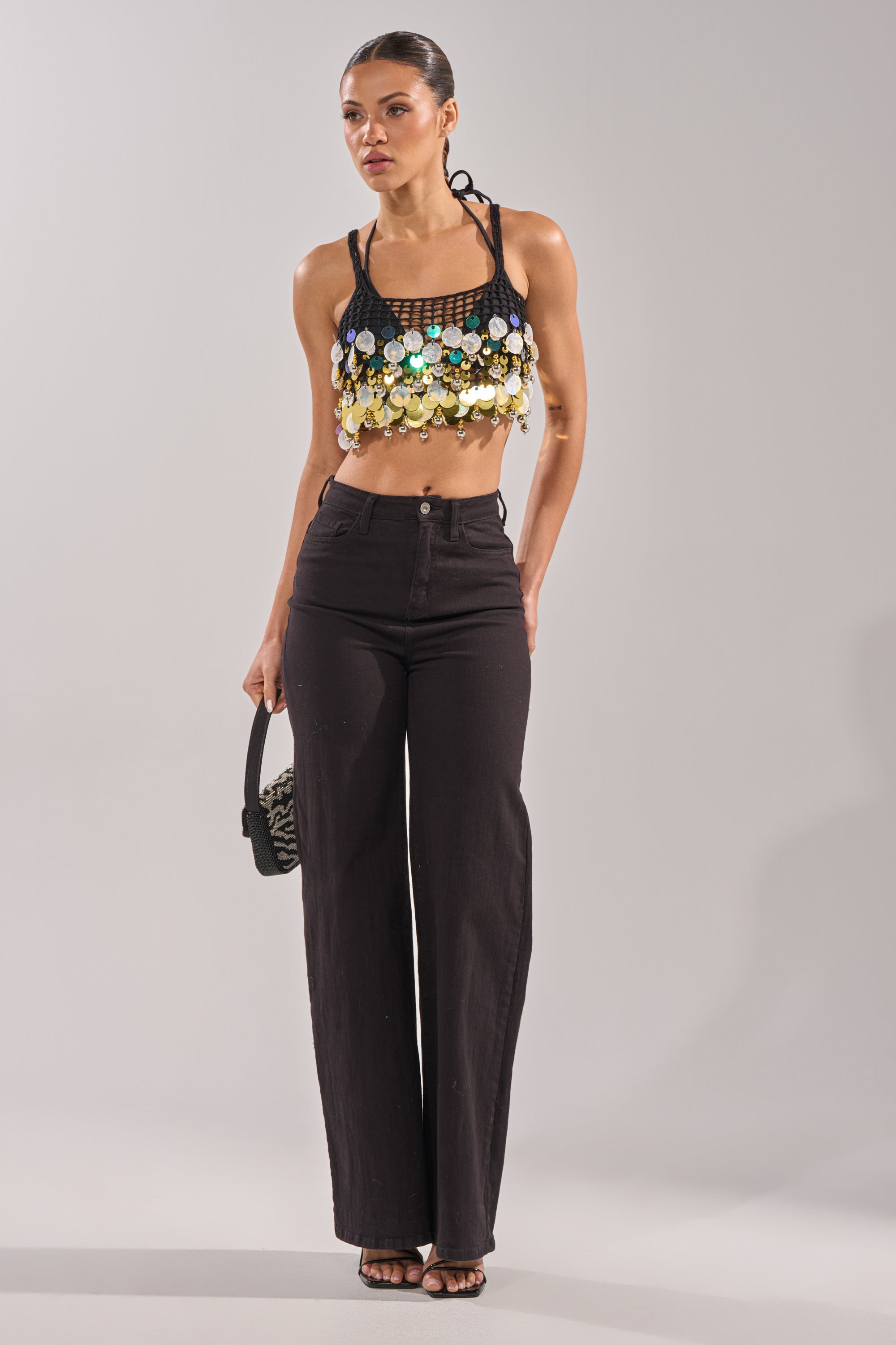 A woman stands in a studio wearing the RESORT LIVING SEQUIN CROCHET TOP, high-waisted black wide-leg pants, strappy heels, and holding a small handbag. She looks to the side with her hair pulled back. 6