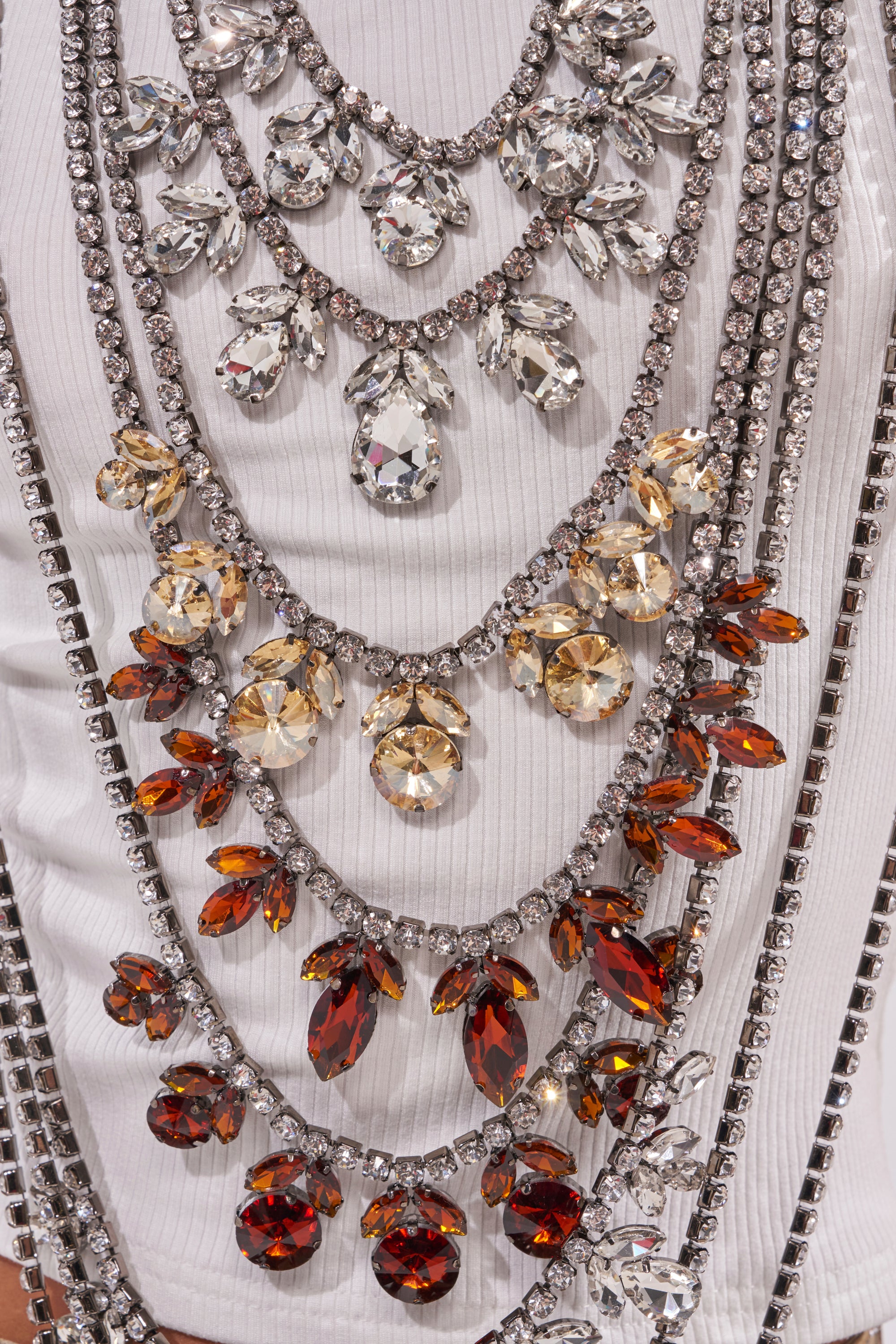Close-up of the CHECK IT OUT EMBELLISHED RHINESTONE TOP, featuring layered statement necklaces with large clear, champagne, and amber crystals, displayed on a white cropped ribbed tank. 9