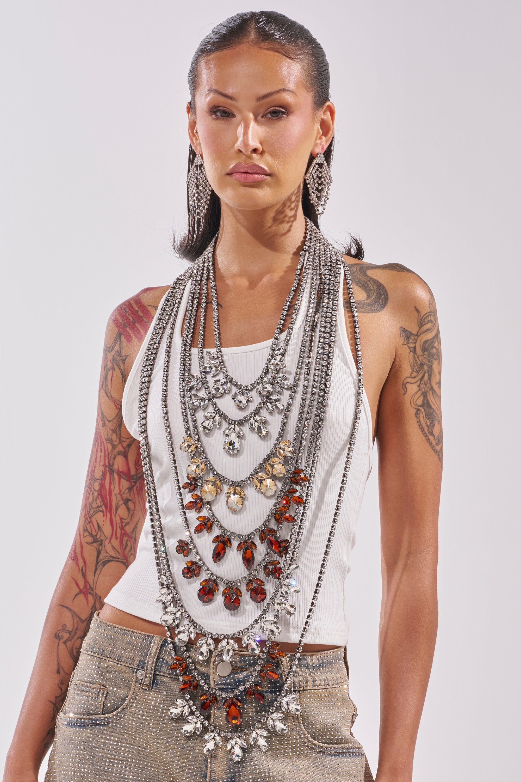A woman with long dark hair, tattoos, and studded beige pants confidently wears the CHECK IT OUT EMBELLISHED RHINESTONE TOP, paired with layered silver and jeweled necklaces and large earrings. 2