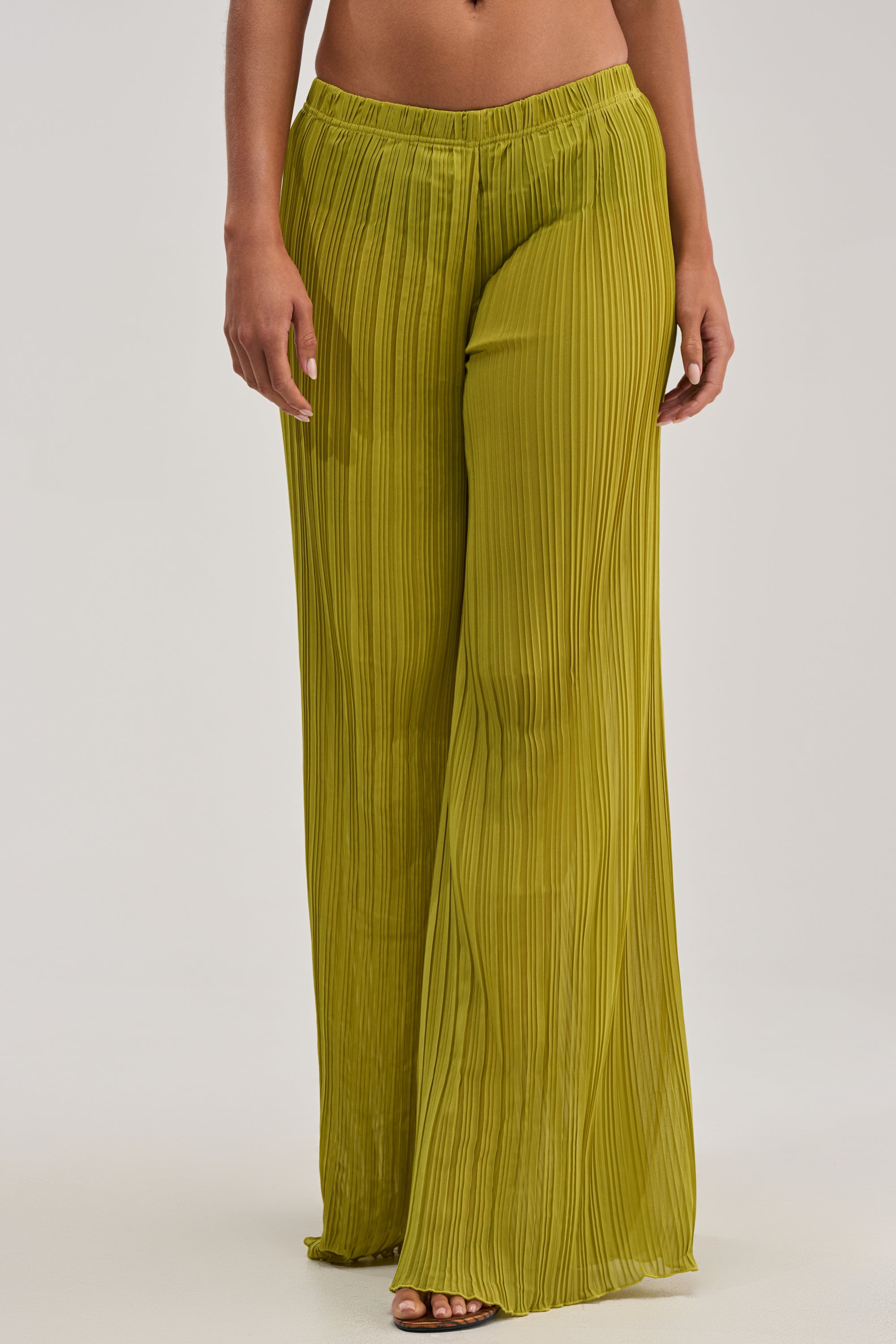A person stands barefoot against a plain light background, modeling the CRUSHIN ON YOU PALAZZO PANT IN CHARTREUSE—high-waisted pleated chiffon pants that showcase effortlessly chic resort style.